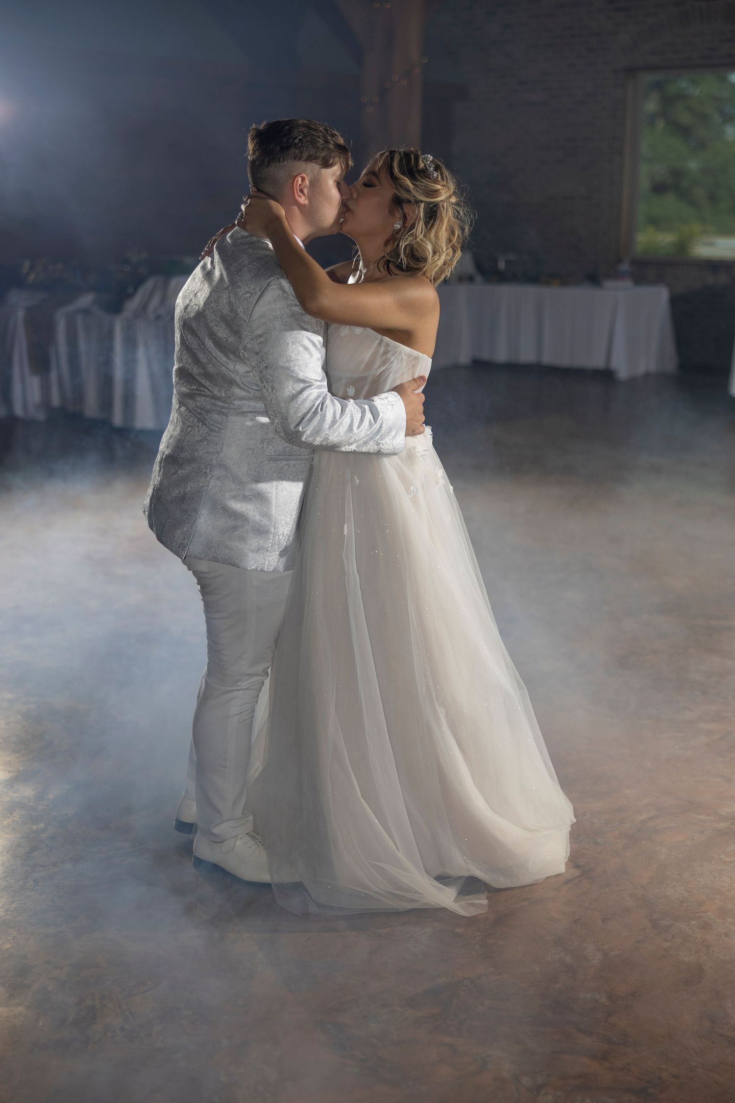 A couple in wedding attire performs their first dance surrounded by atmospheric fog on a dark dance floor.