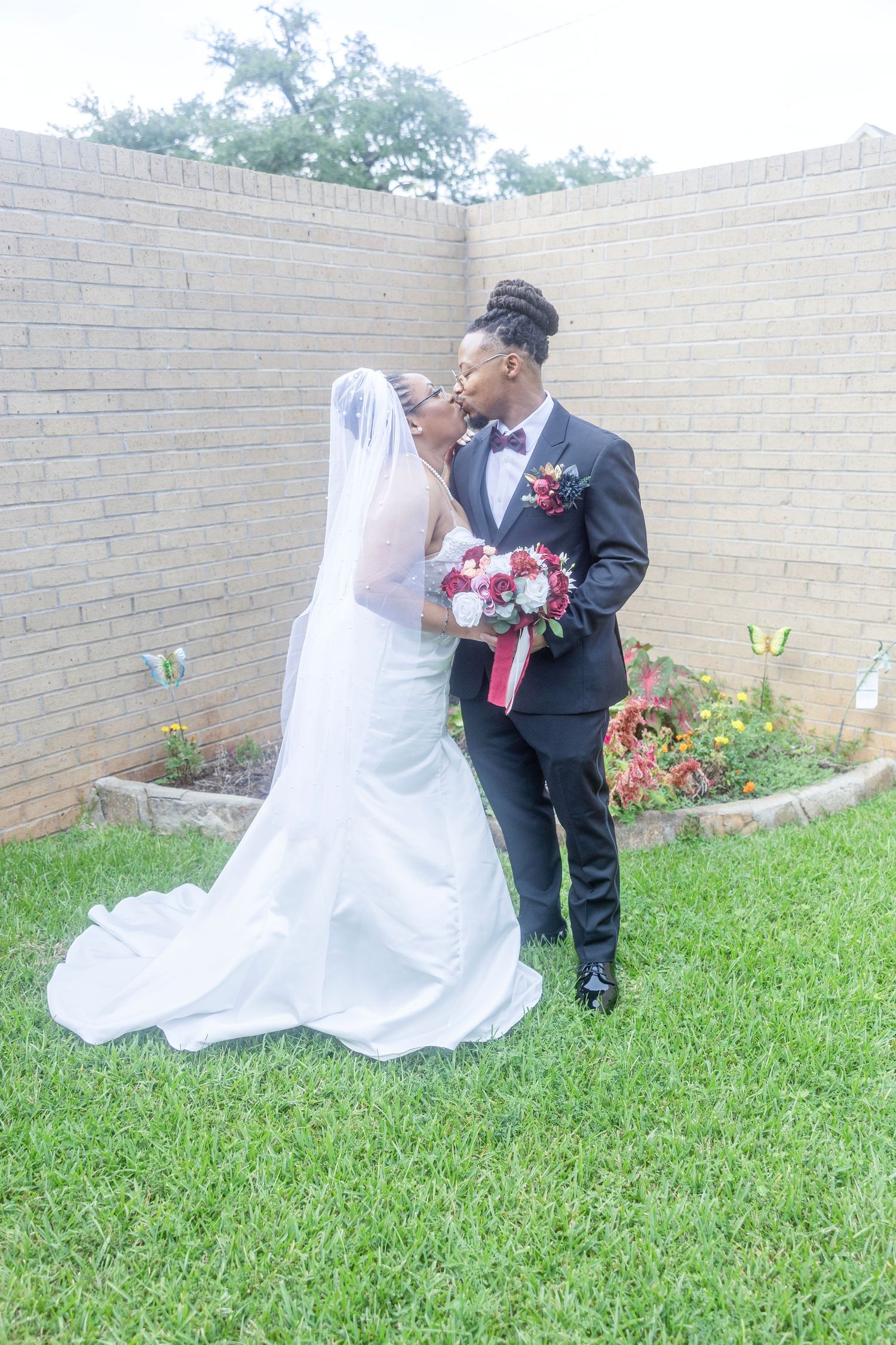 A couple in wedding attire shares romantic moments on a grassy lawn beside a brick wall.