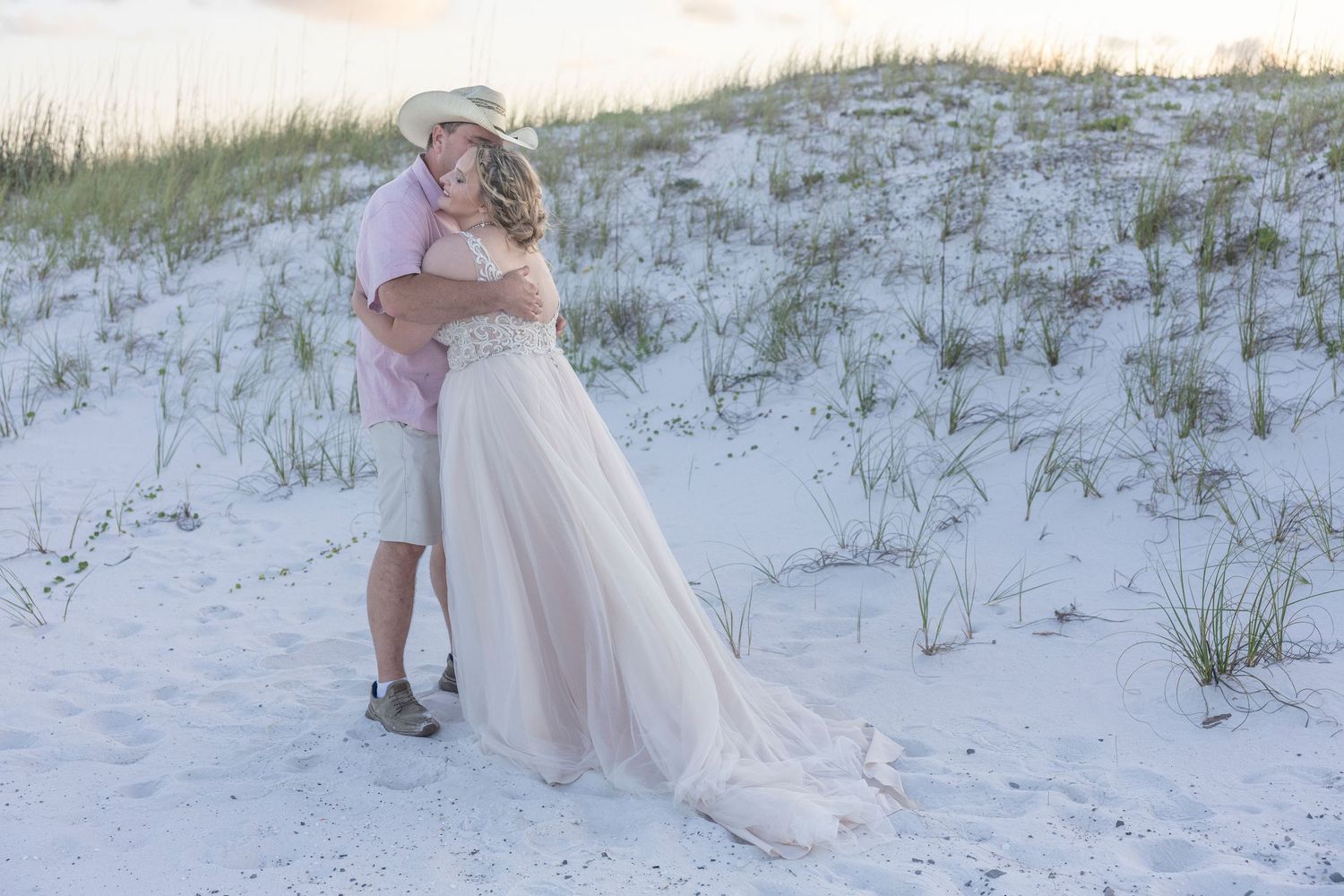 A couple shares an intimate moment on a sandy beach with sea oats and dunes at sunset.