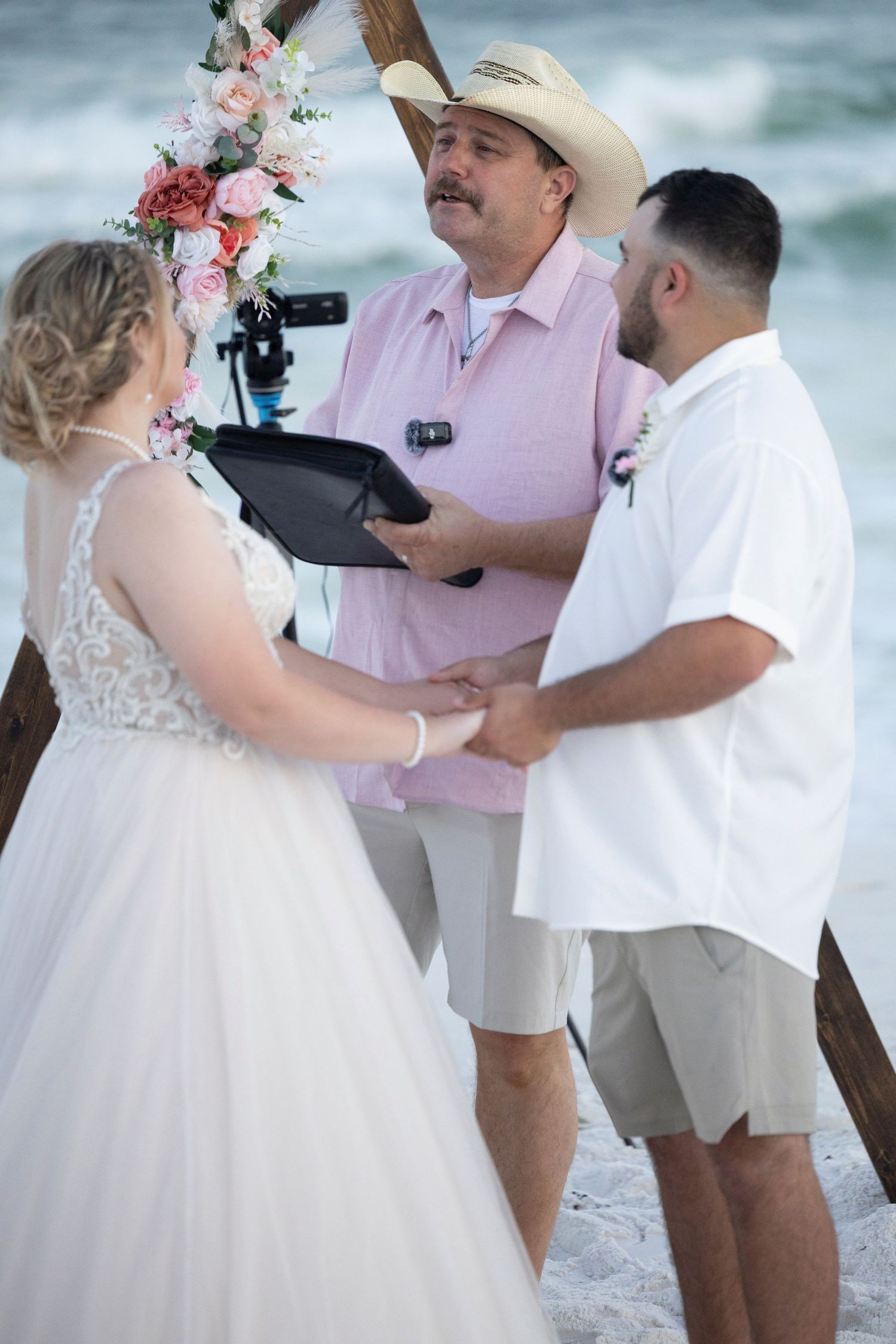 A beachside wedding ceremony takes place with people in white and pink attire against an ocean backdrop.
