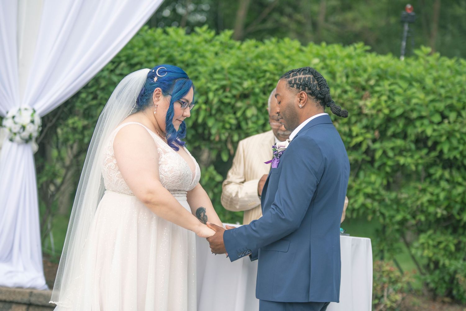 A couple shares an intimate moment under a white draped wedding arch surrounded by lush green foliage.