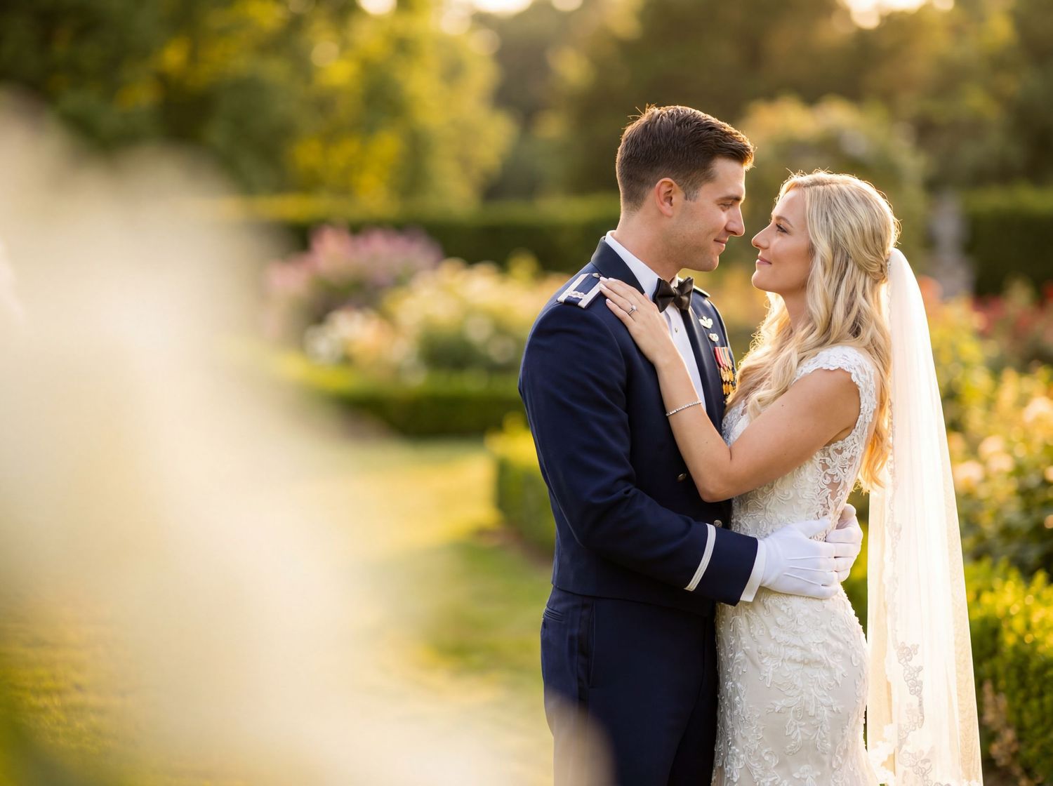 Bride and groom share a romantic embrace in a sunlit garden, with the groom in military dress blues.
