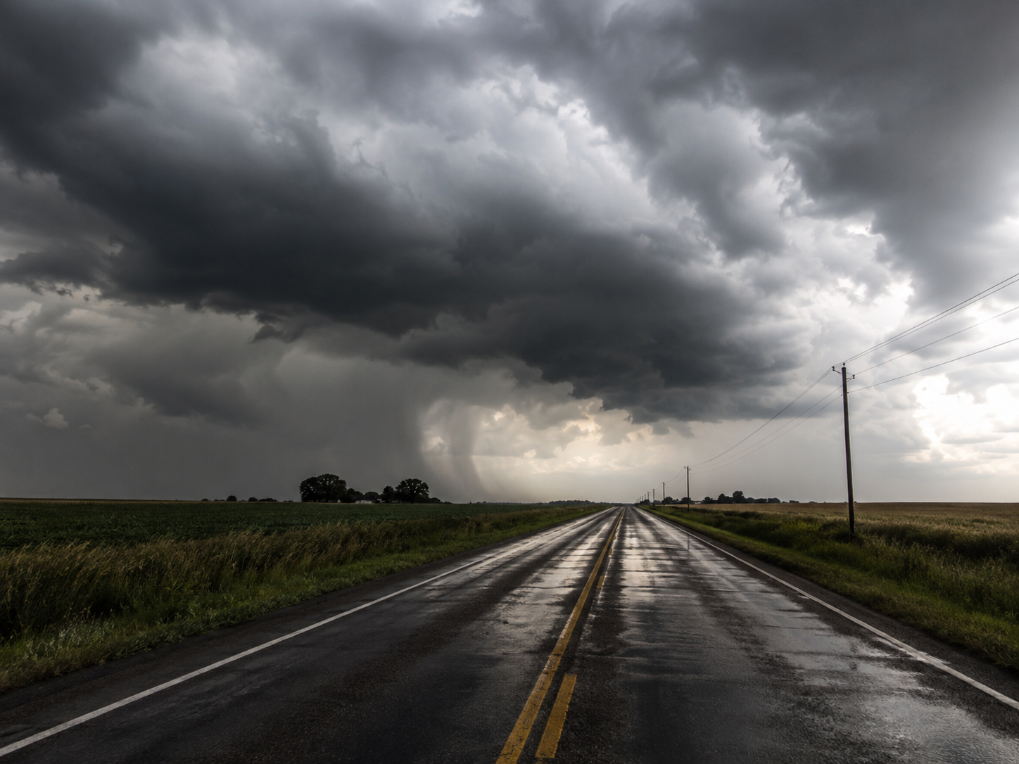 Wet rural road stretching toward dramatic storm clouds over flat farmland.