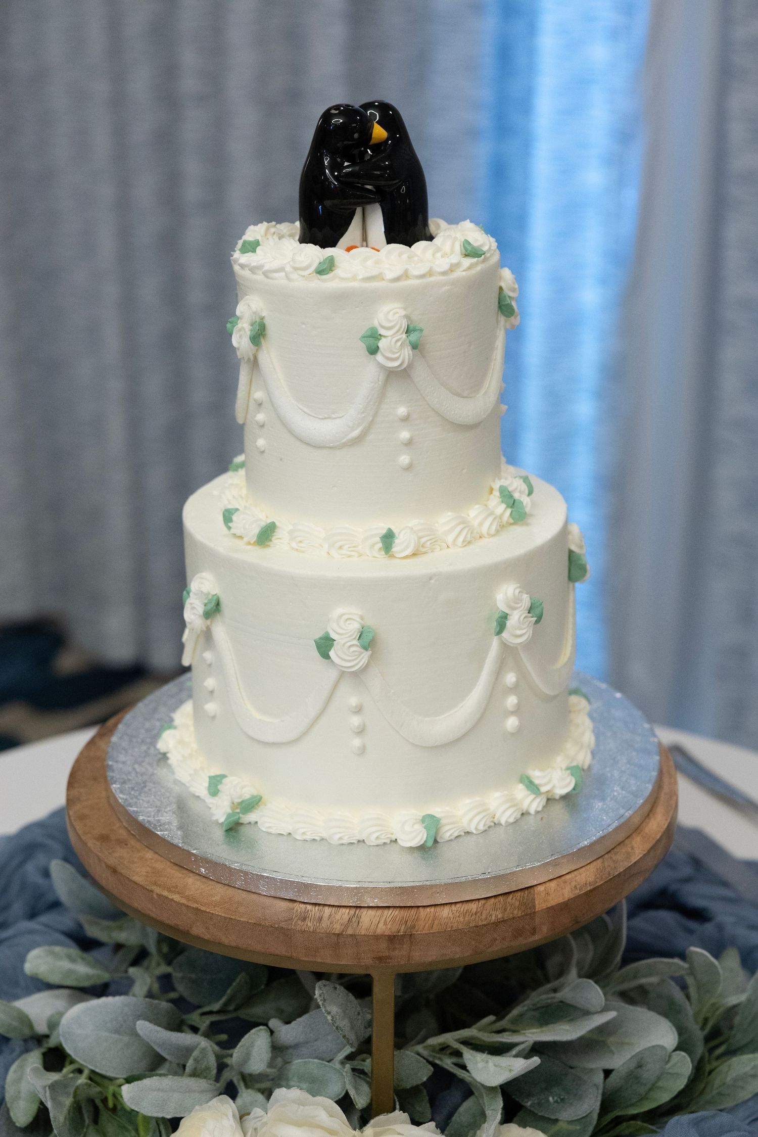 Three-tier white wedding cake with mint green floral accents and black penguin cake toppers on a wooden stand.
