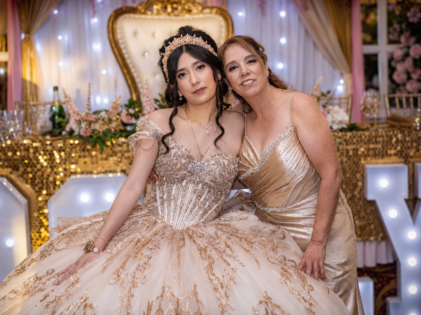 Two women in elegant gold gowns pose together at a quinceañera celebration with festive decorations.