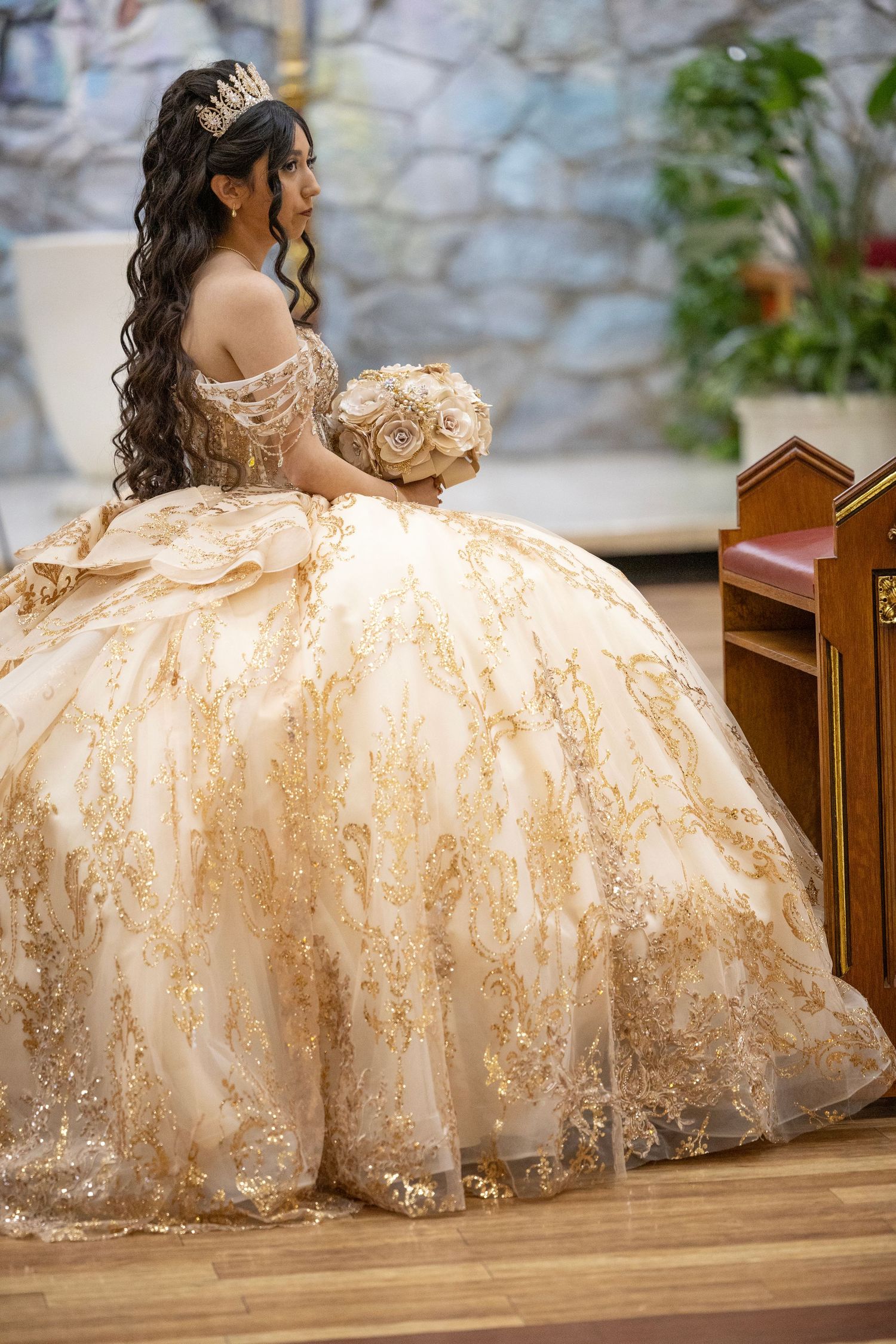 Young woman in elegant gold quinceañera ball gown with tiara holding floral bouquet in a church setting.