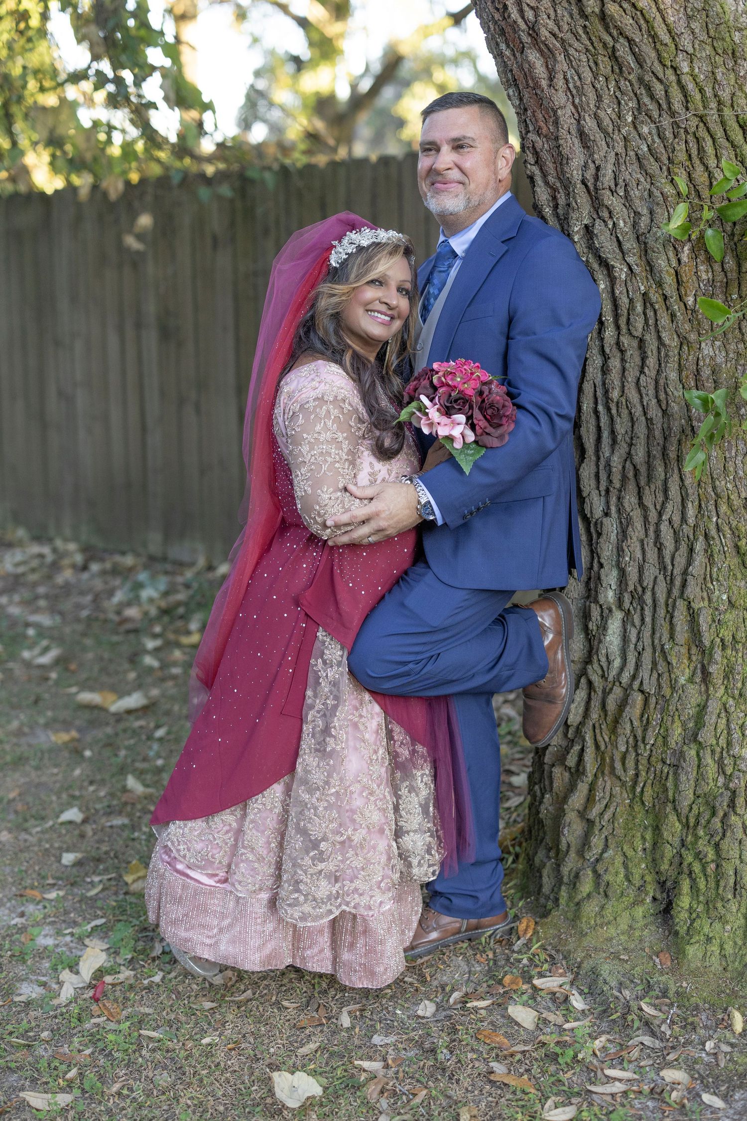 A couple poses together by a tree trunk, with the bride wearing a pink dress and burgundy veil holding a floral bouquet.
