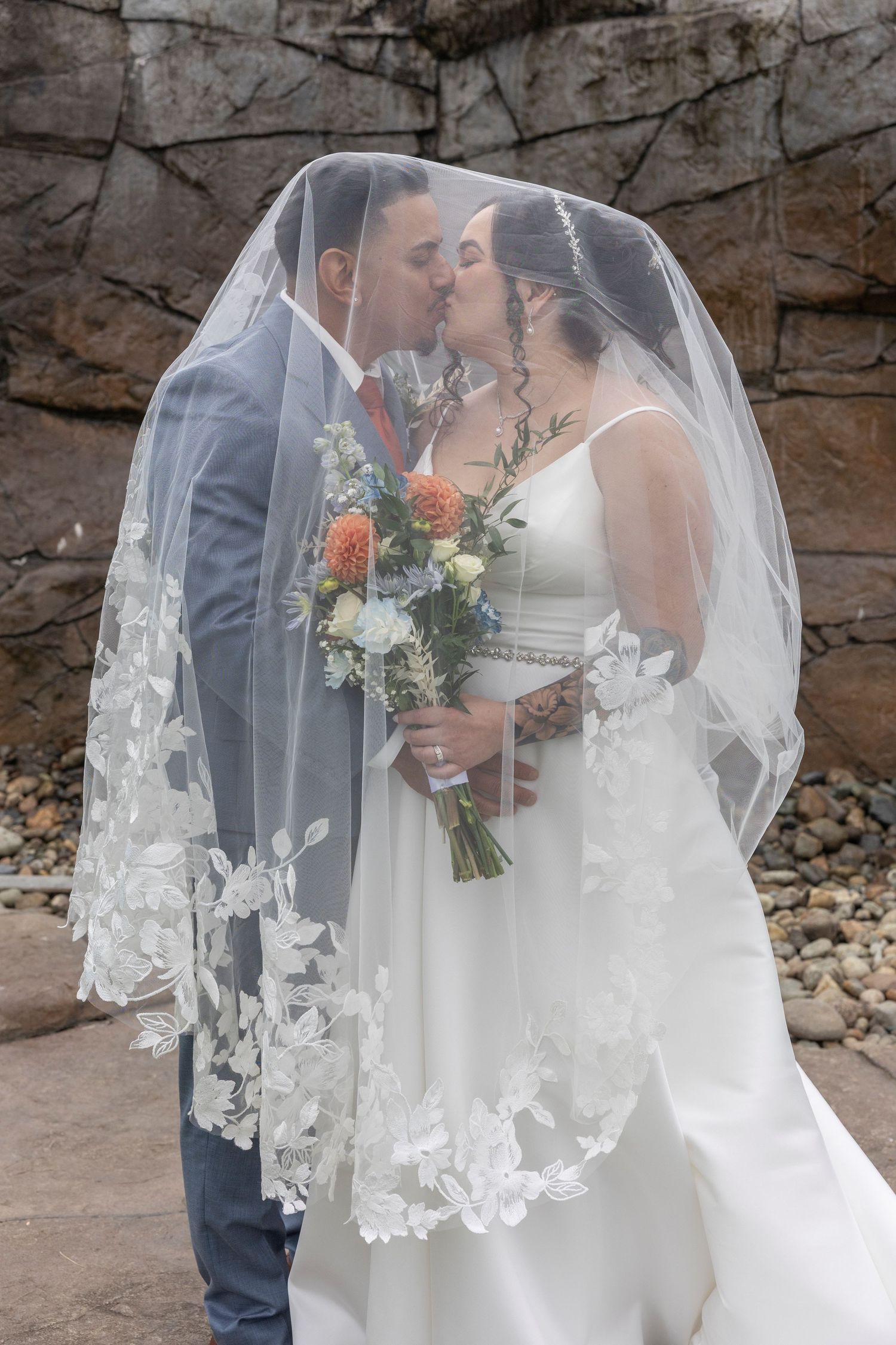 A romantic wedding couple shares an intimate moment against a stone wall backdrop while the bride wears a flowing lace veil.