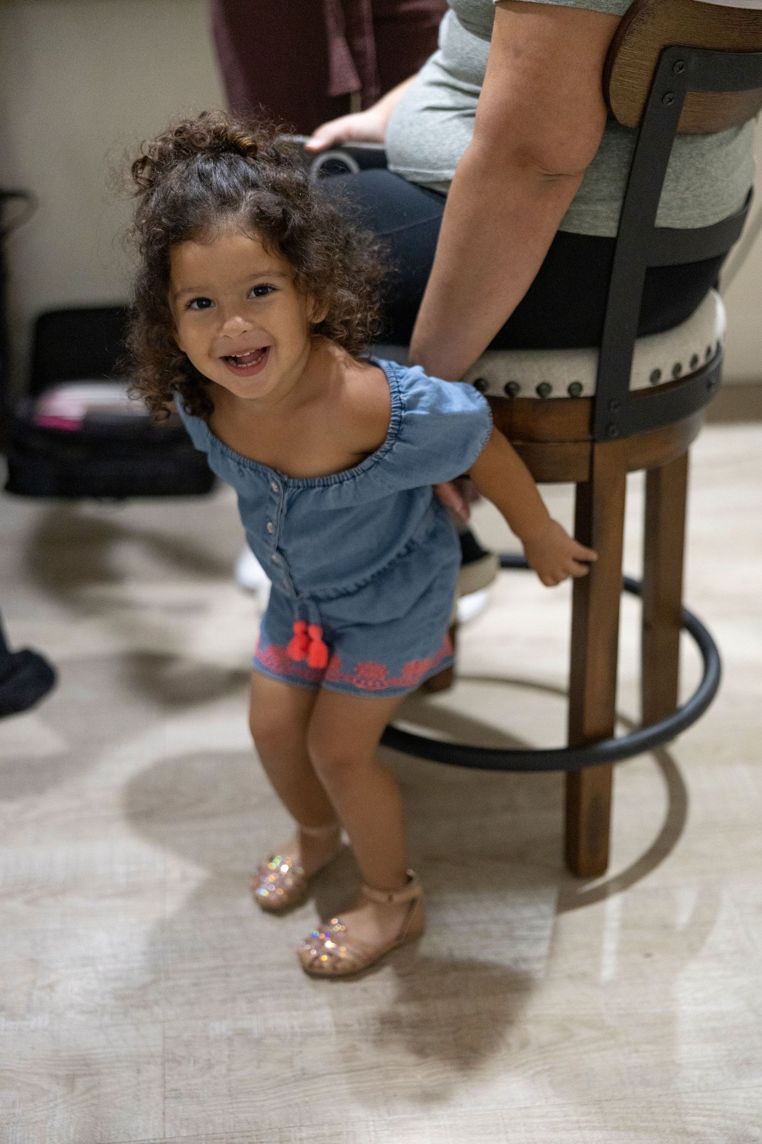 A small child wearing blue denim shorts and sandals stands near a wooden chair on a light-colored floor.