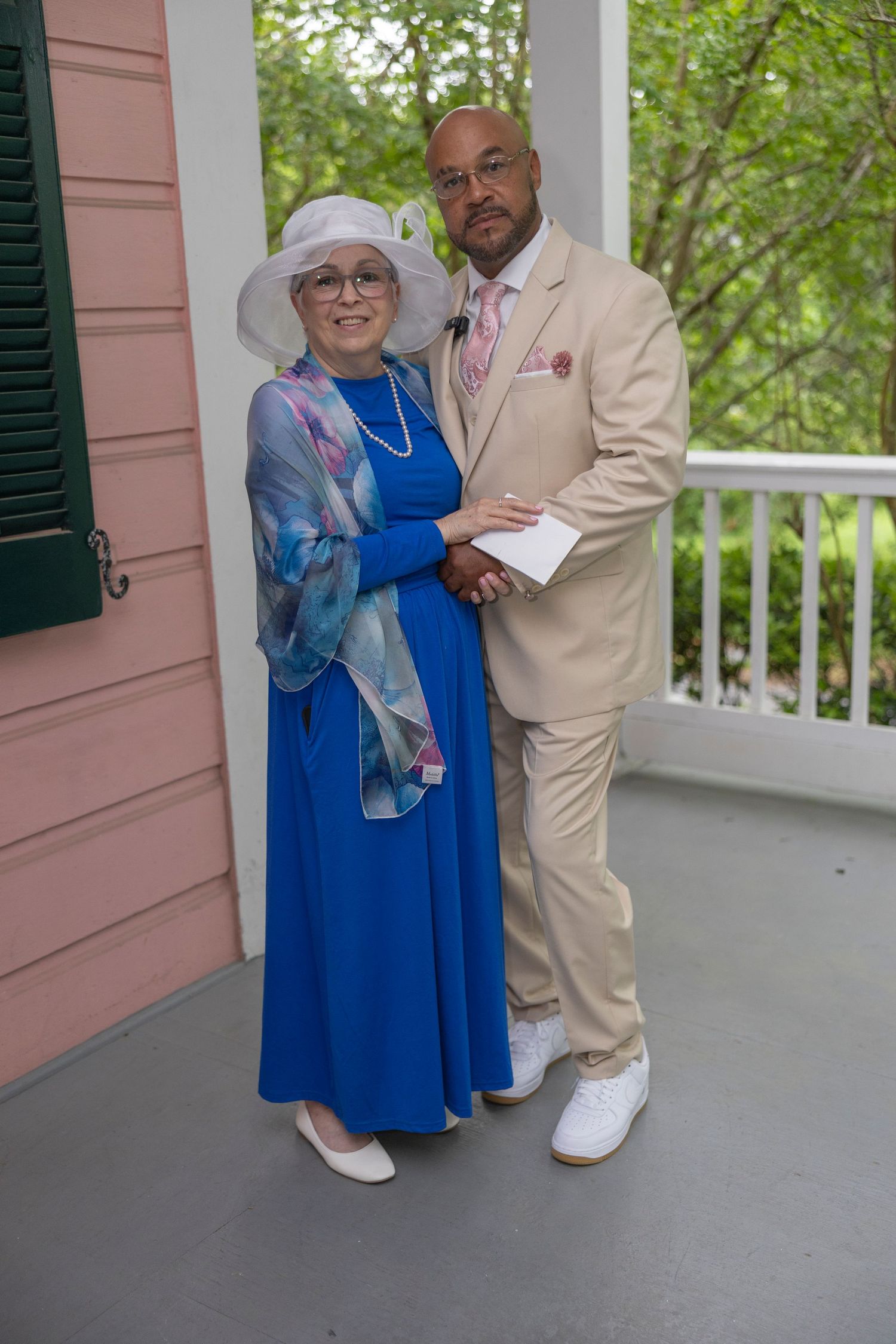 A couple poses together on a porch, with the woman in a blue dress and white hat, and the man in a beige suit.