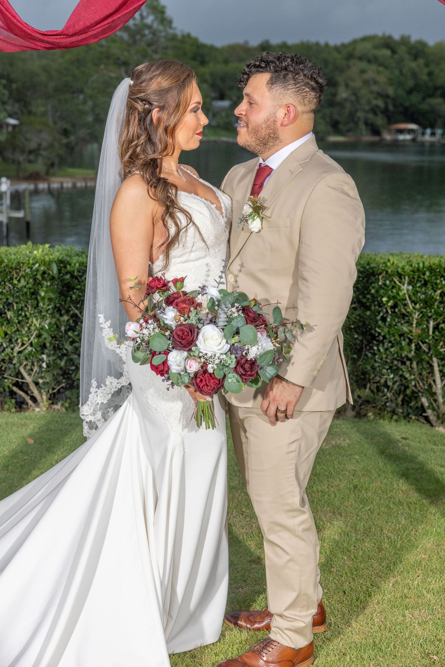 A newlywed couple poses outdoors by a lake, with the bride holding a red and white bouquet and wearing a satin gown.