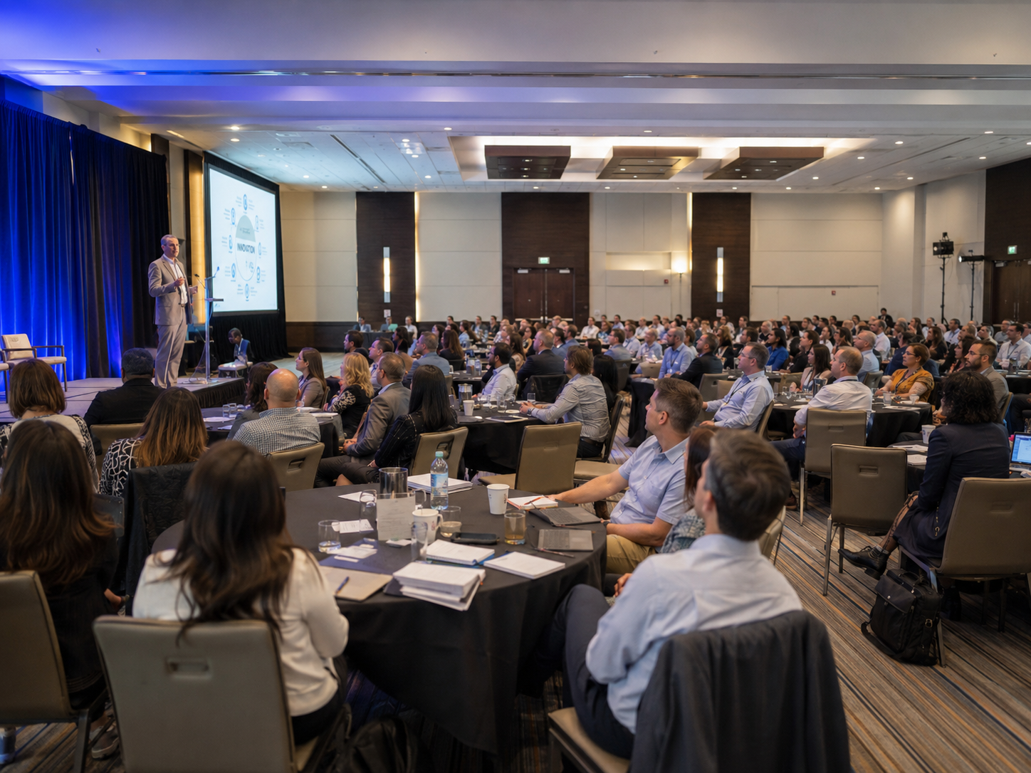 Large audience of professionals seated at round tables during a keynote presentation at a conference.