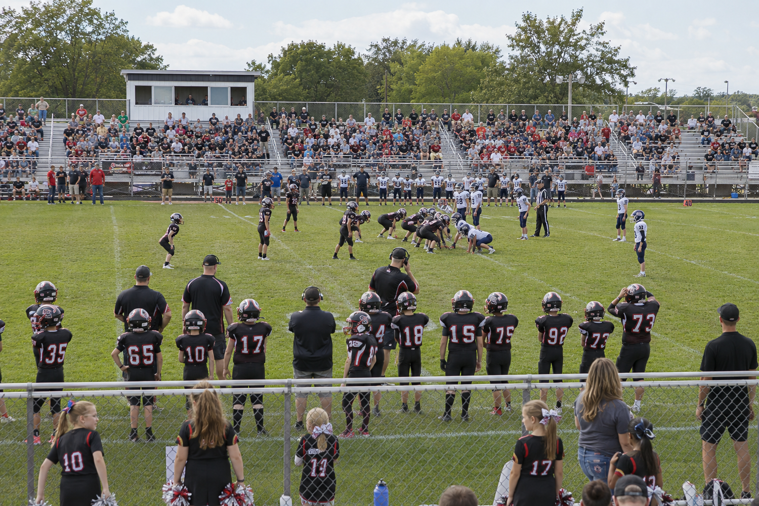 High school football game with players on field and packed bleachers of spectators on a sunny day.