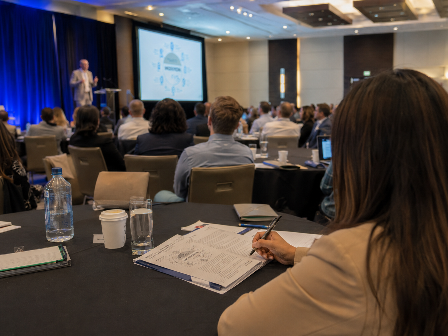 Attendee taking notes during a business conference presentation in a modern hotel meeting room.