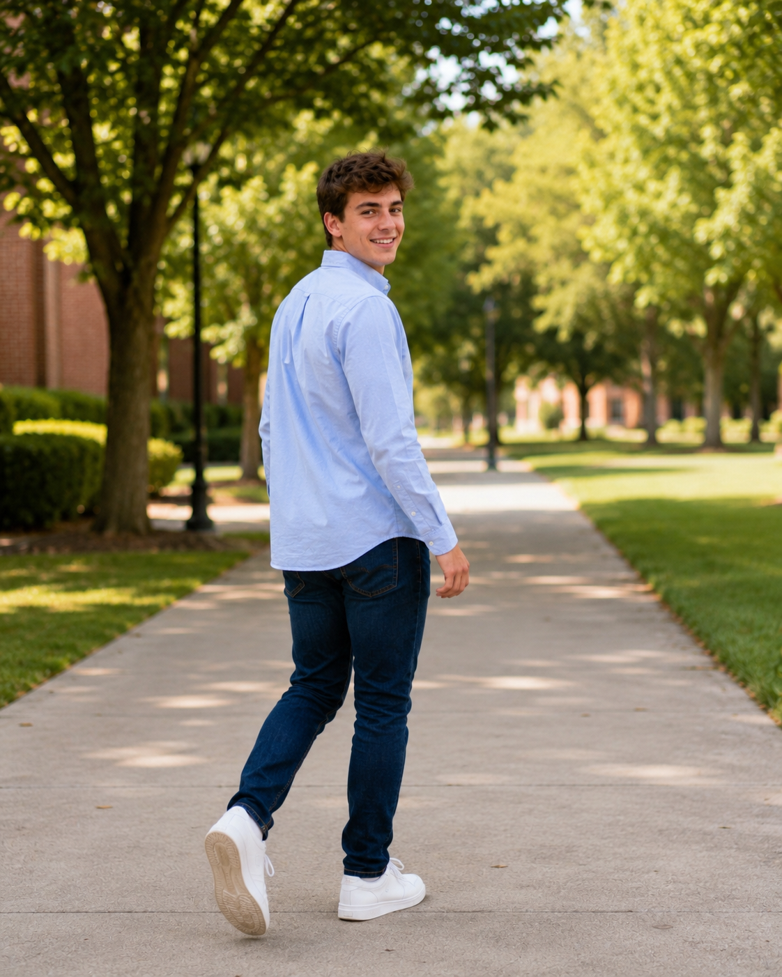 Young man in blue shirt and navy jeans walks along a tree-lined campus path, smiling over his shoulder.