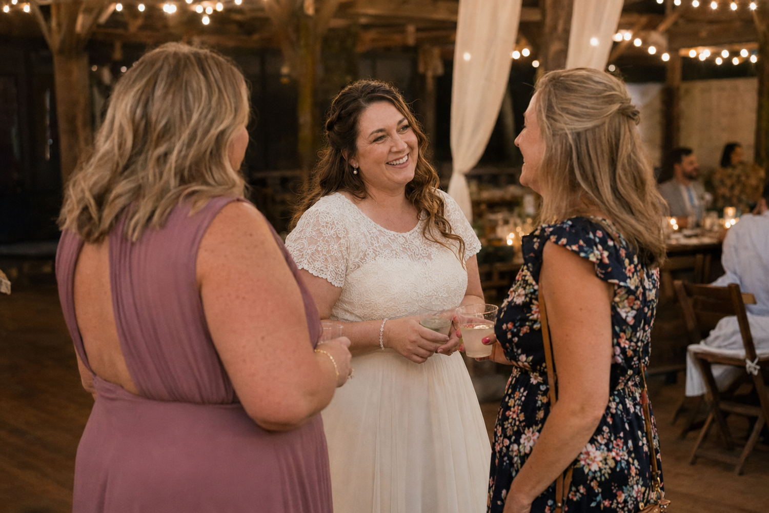 Bride in lace wedding dress laughing with two guests inside a rustic barn wedding venue.