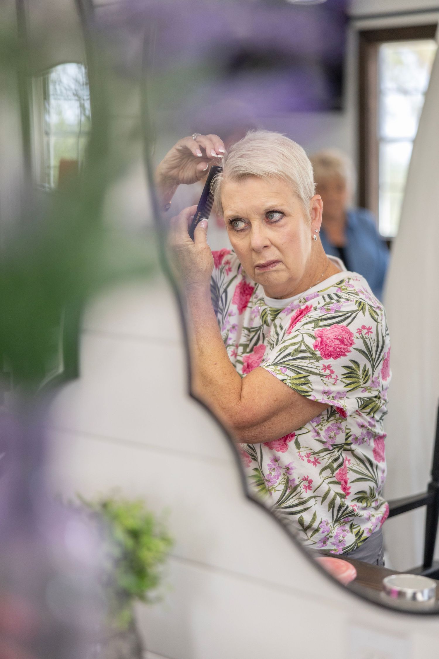 Older woman with short gray hair brushing her hair while looking in a mirror, wearing a floral pink top.