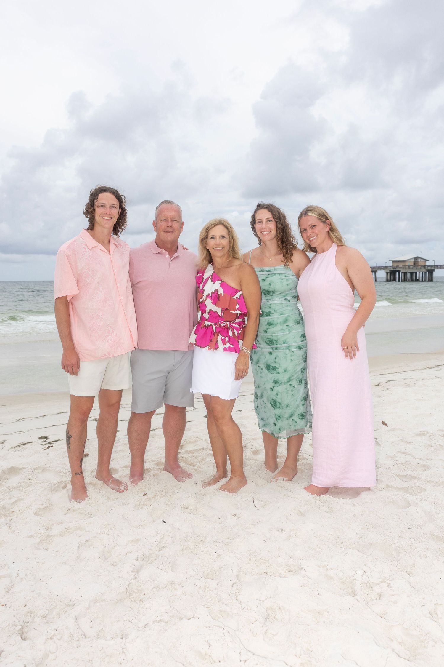 A family group poses together on a white sandy beach wearing pastel colors with a pier visible in the background.