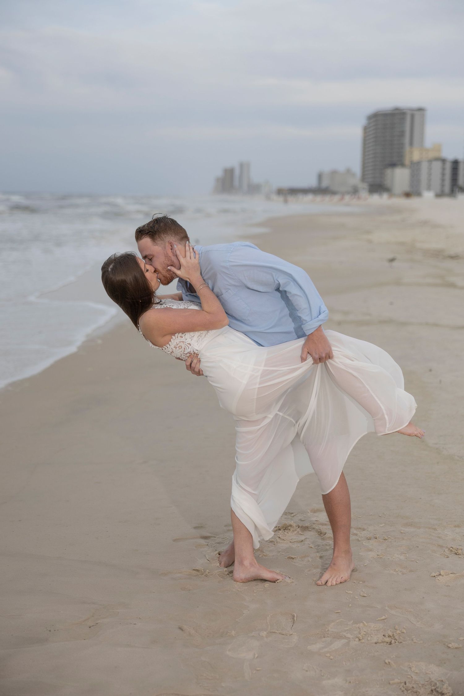 Couple sharing a romantic kiss on the beach, woman in white dress and man in blue shirt, with ocean waves in the background.