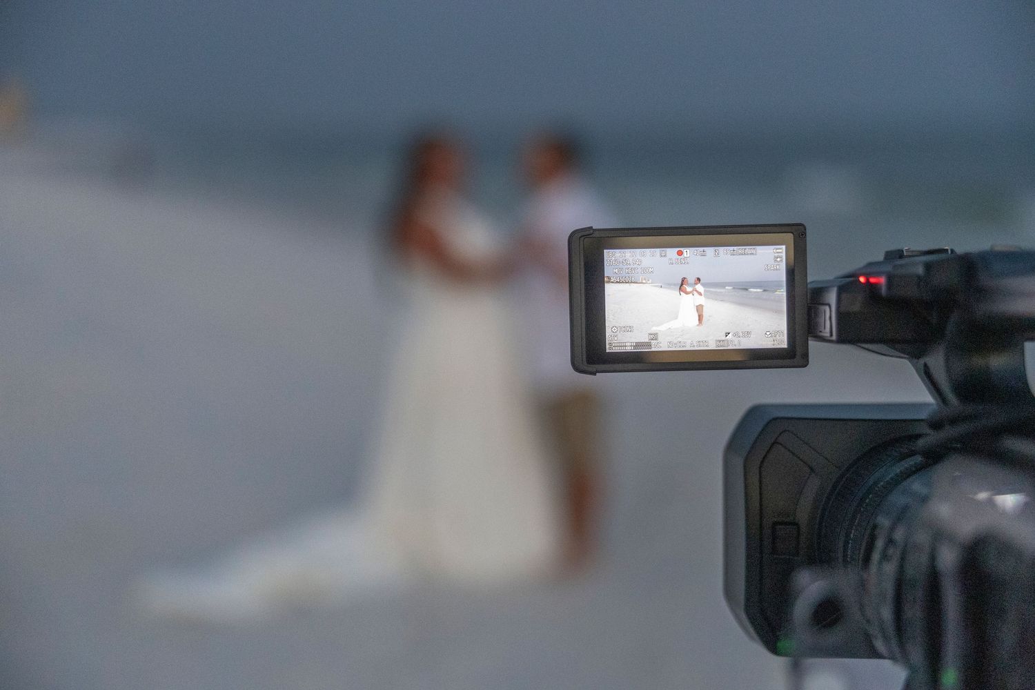 A video camera LCD screen displays a wedding ceremony being filmed on a beach with blurred figures in the background.