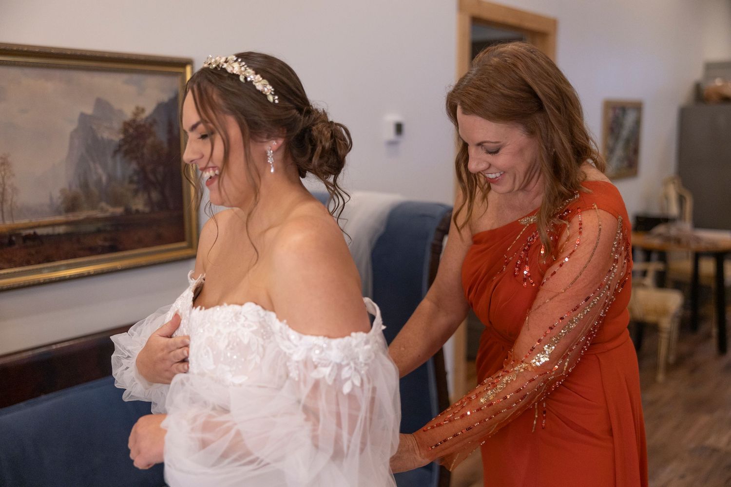 Two women share an emotional moment as one helps adjust a white off-shoulder wedding dress in an indoor setting.