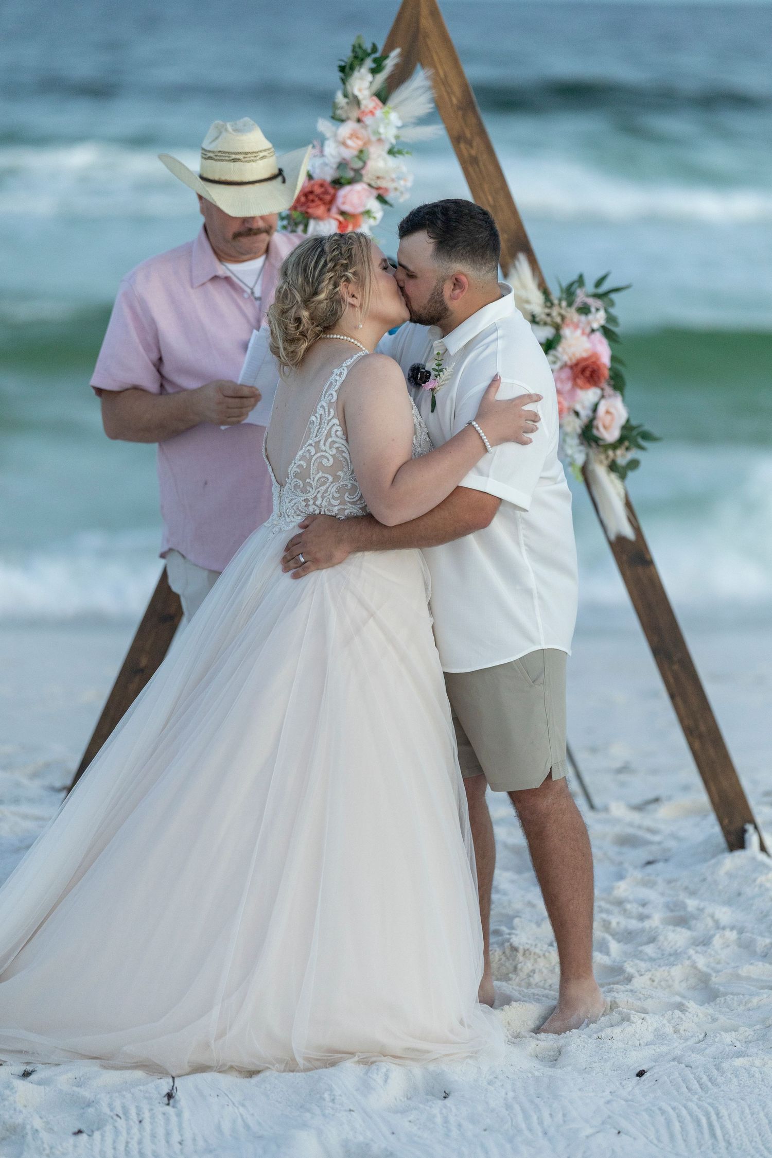 A couple shares a romantic kiss on a white sandy beach during their beachside wedding ceremony.