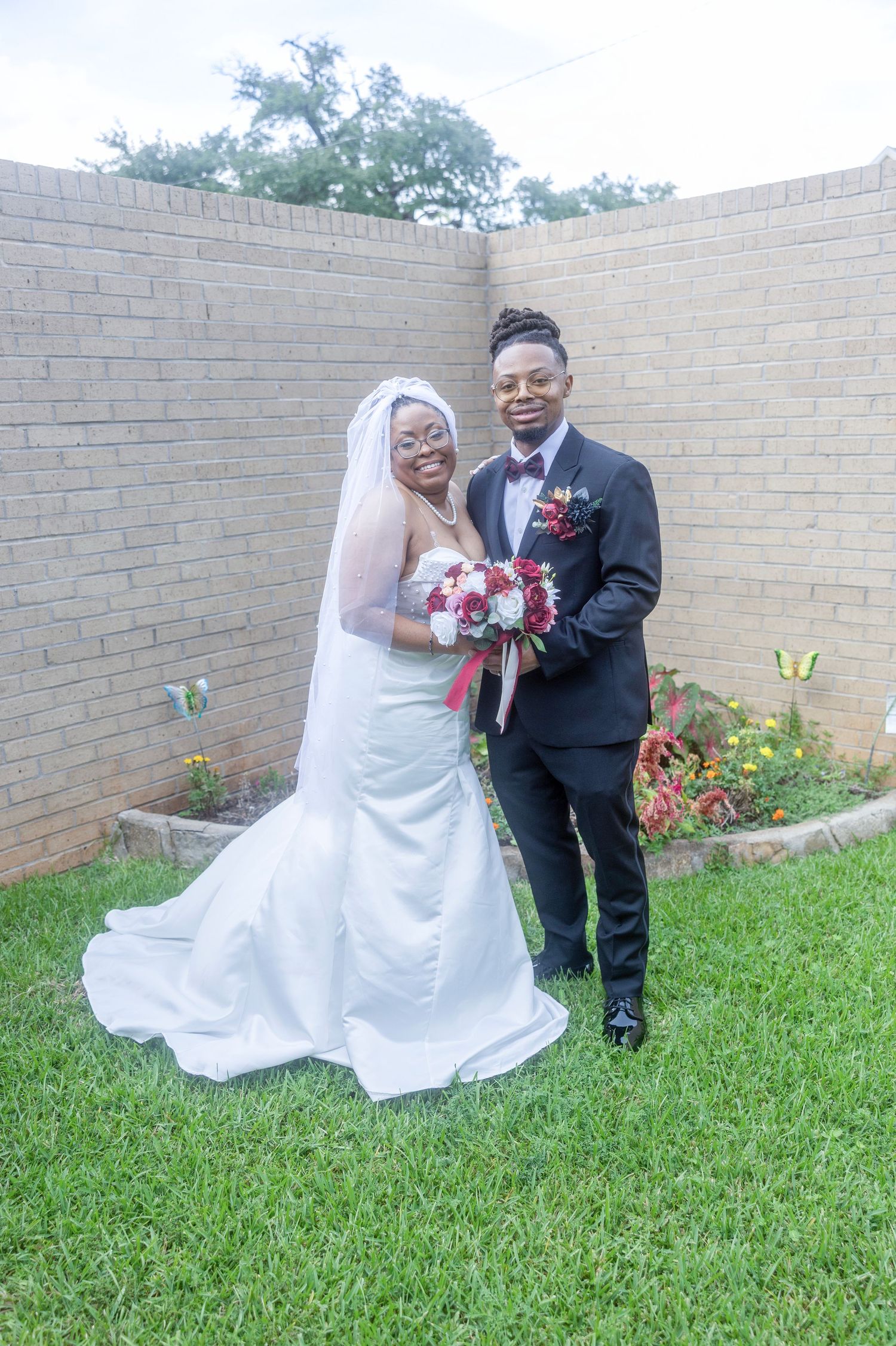 A happy couple shares joyful moments in a garden setting against a brick wall with the bride wearing a flowing white dress.