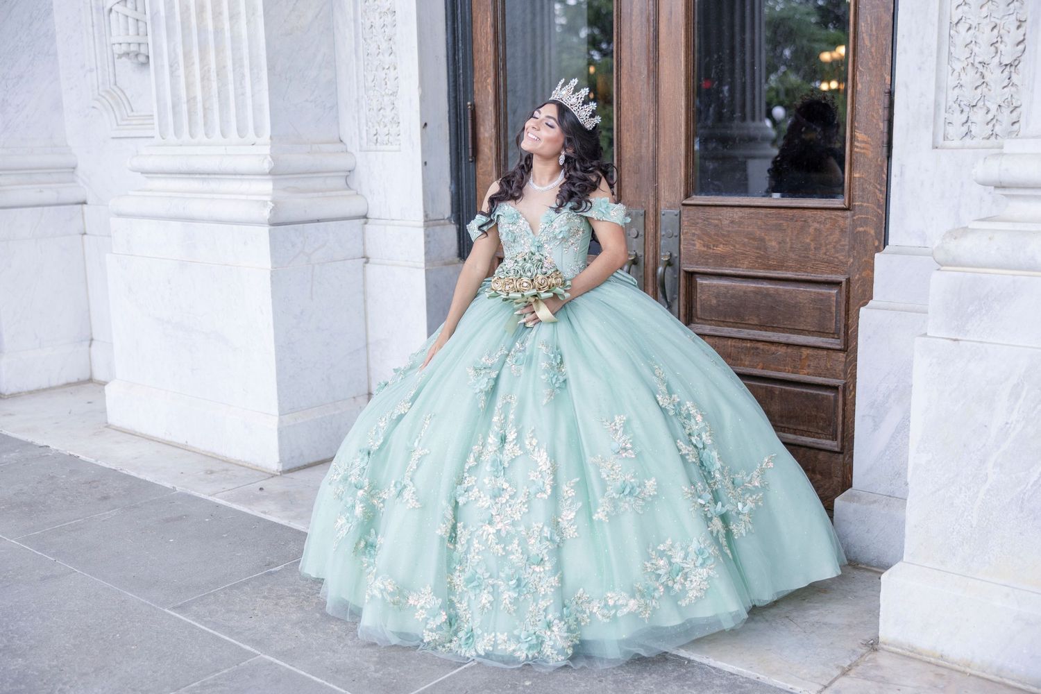 A person wearing an elegant mint green ball gown with white lace appliques poses in front of ornate white architecture.