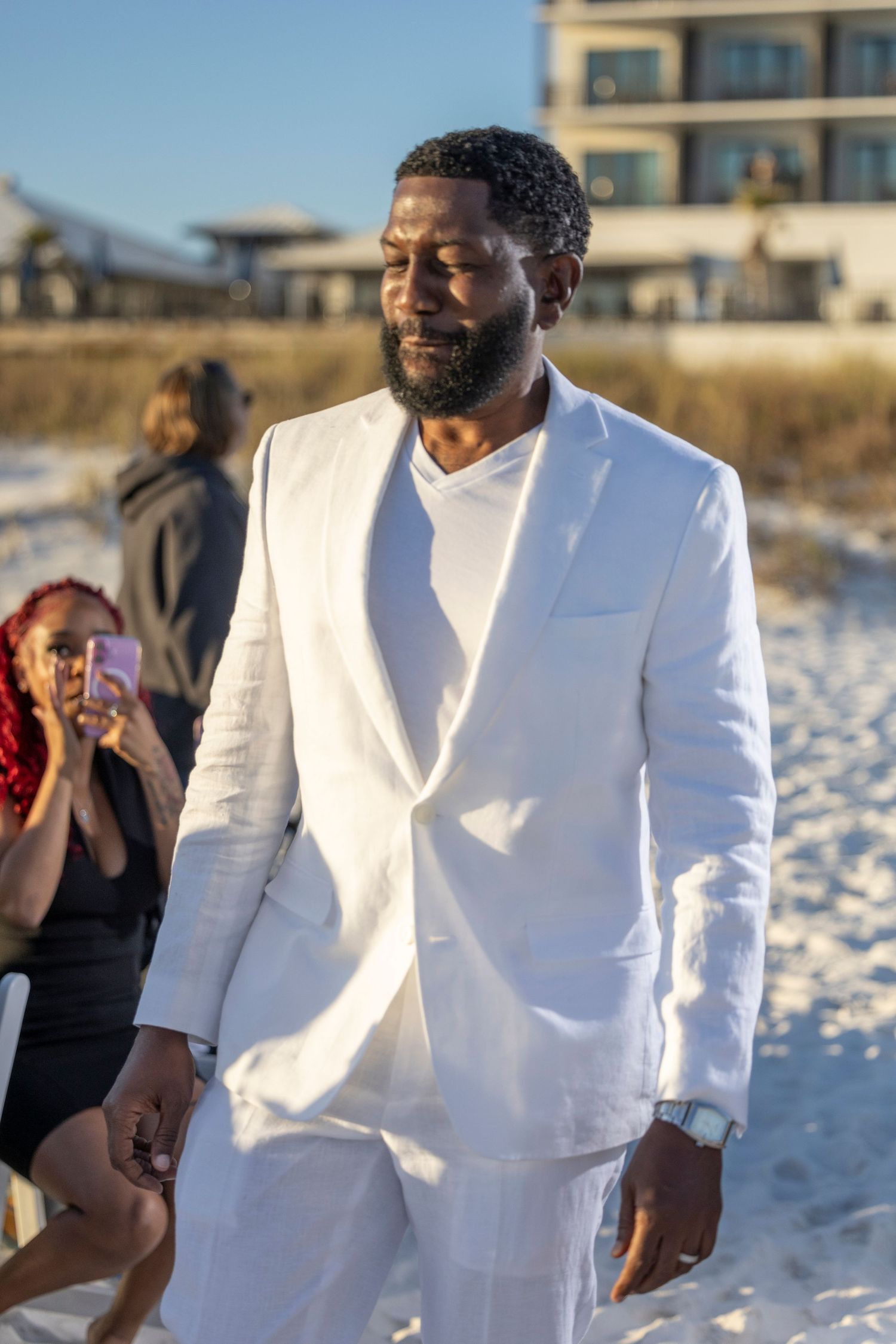 A person in a white linen suit smiles while walking on a sunny beach during sunset.