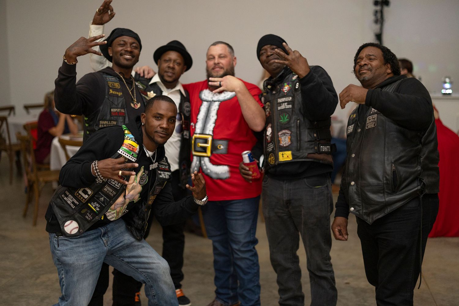 Group of motorcycle club members wearing leather vests and jackets gather together in a casual setting.