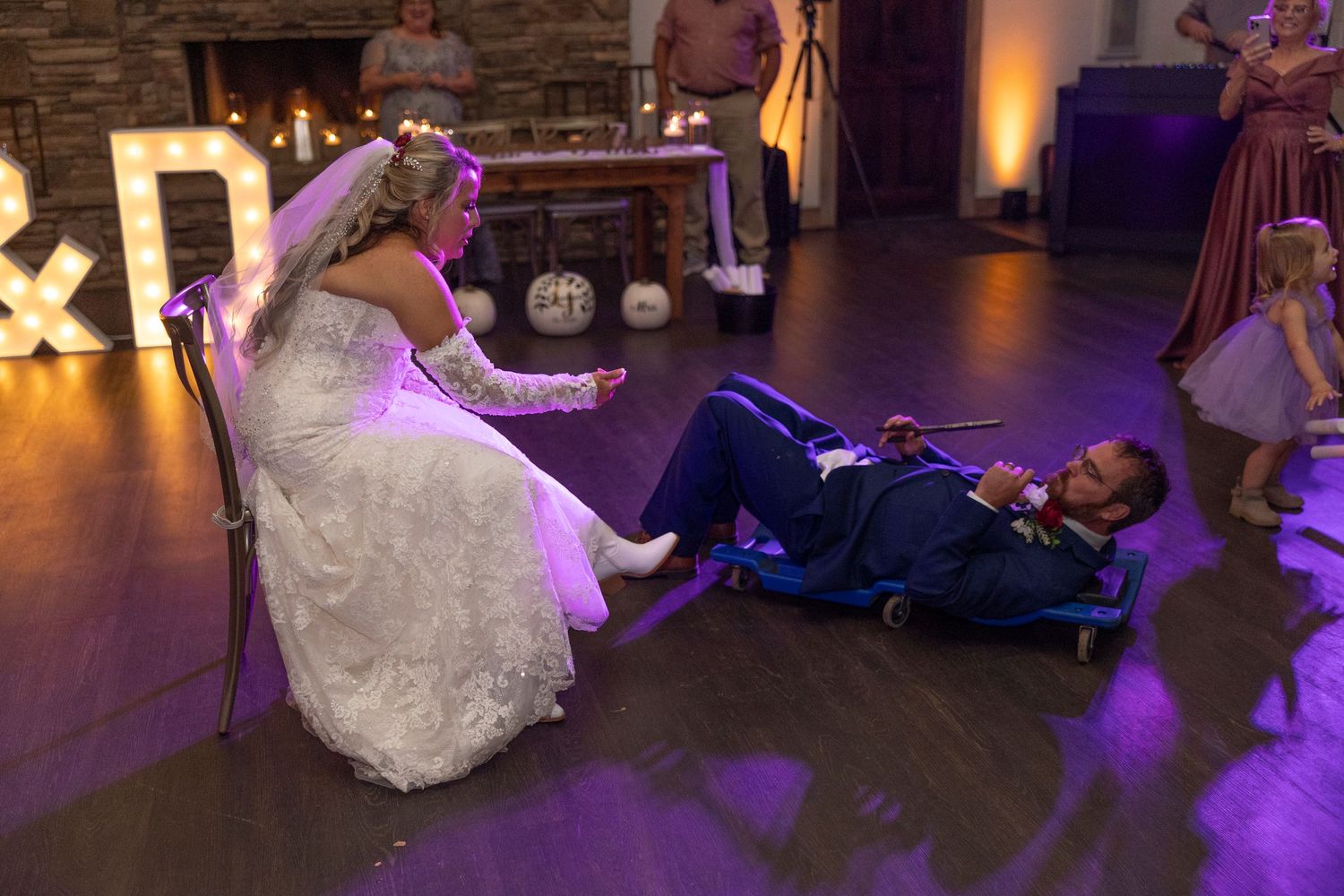 Wedding guests dance together on a dimly lit dance floor with purple lighting during an evening reception celebration.