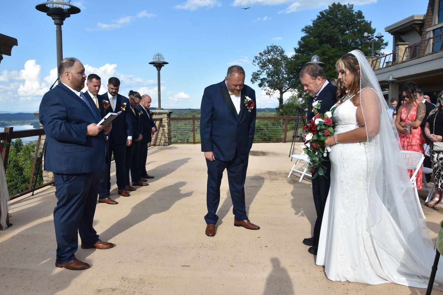 A wedding party poses together on a scenic outdoor terrace overlooking a beautiful landscape on a sunny day.