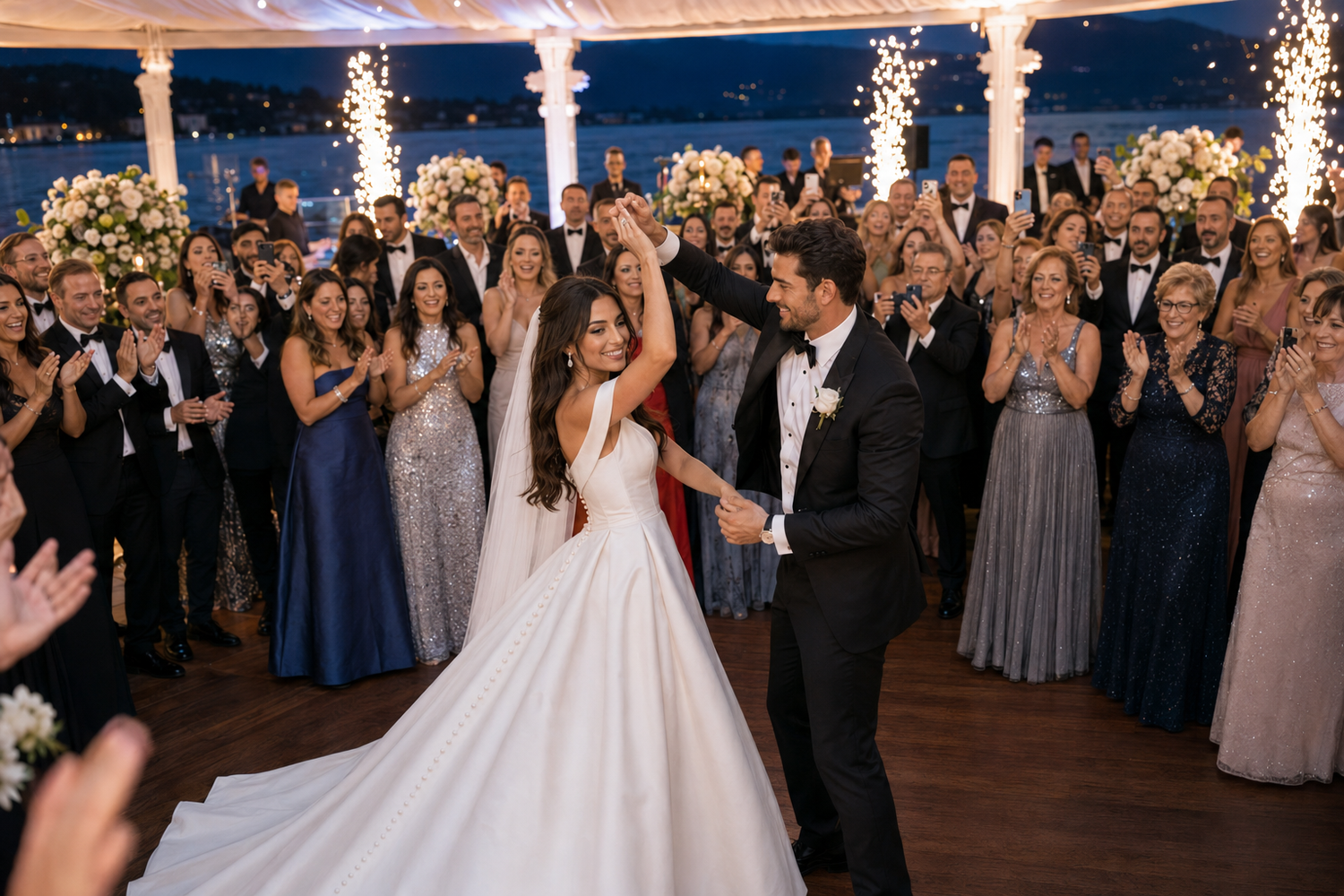 Bride and groom share first dance surrounded by cheering guests with sparklers at elegant waterfront wedding reception.