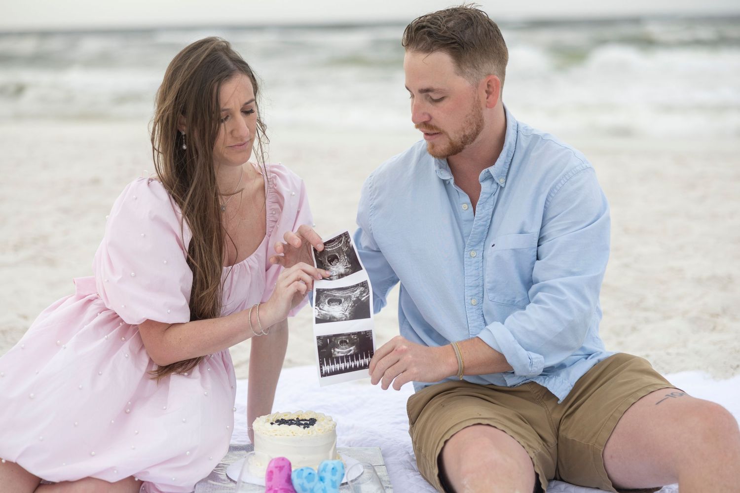 Couple sitting on beach blanket examining ultrasound photos during a maternity announcement photoshoot.