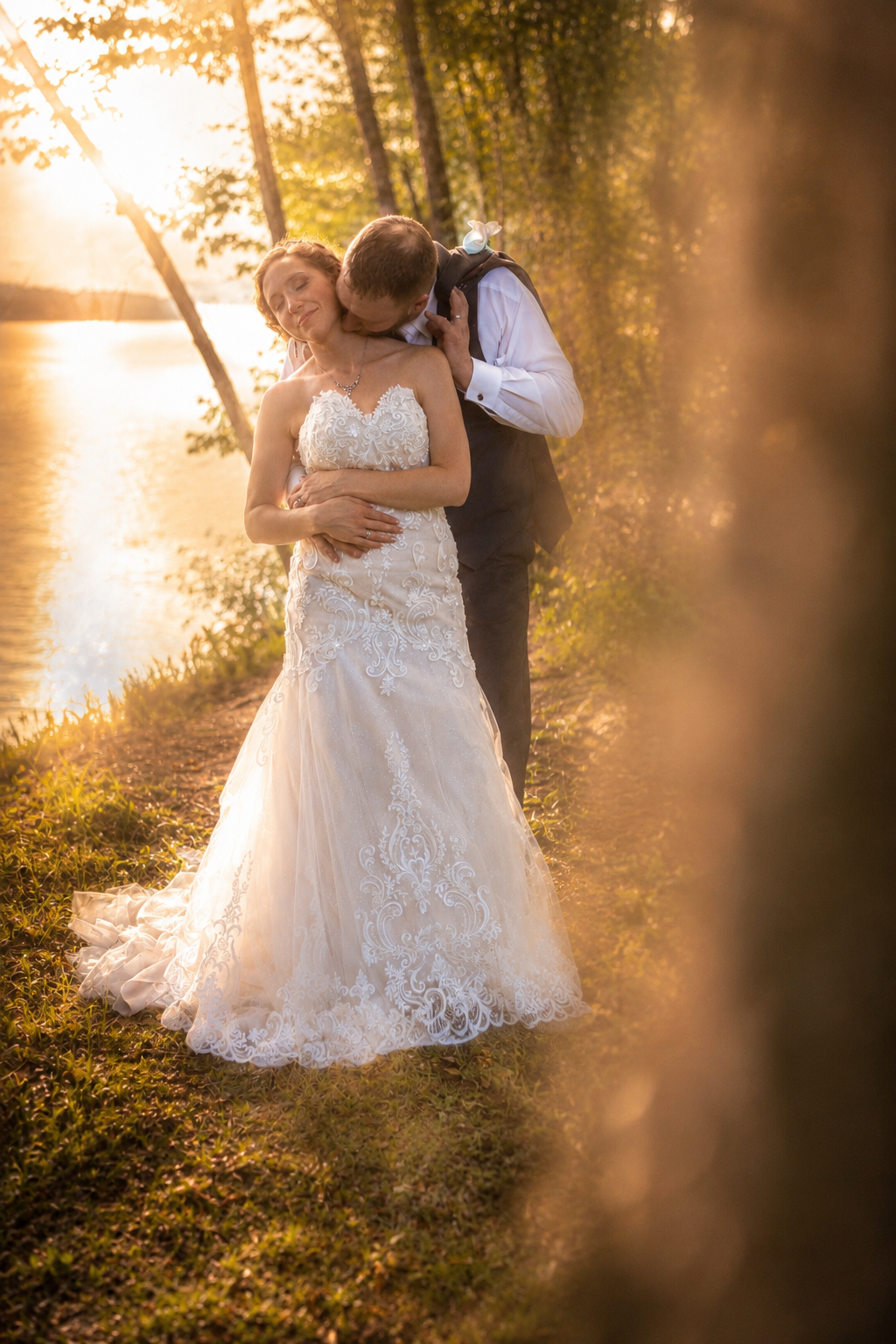 Bride and groom embrace at golden sunset by a lake, she wears a lace mermaid gown amid glowing trees.