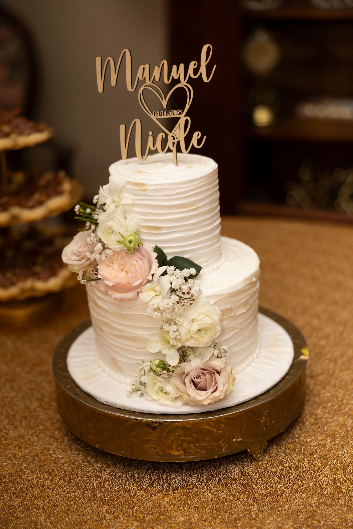 Elegant two-tier white wedding cake with blush roses and gold name topper on metallic gold cake stand.