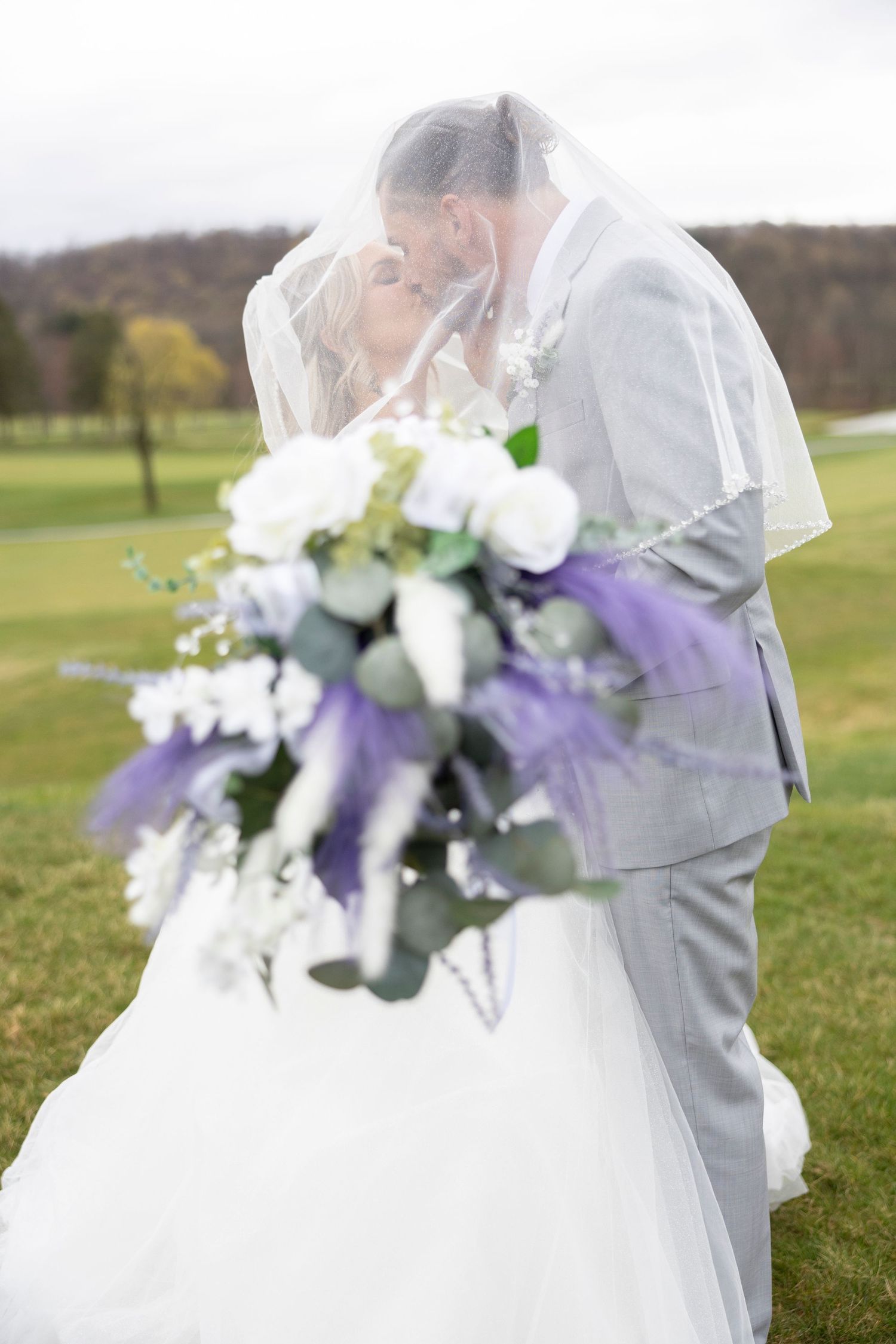 Newlywed couple embraces under a veil on a golf course with a beautiful purple and white floral bouquet.