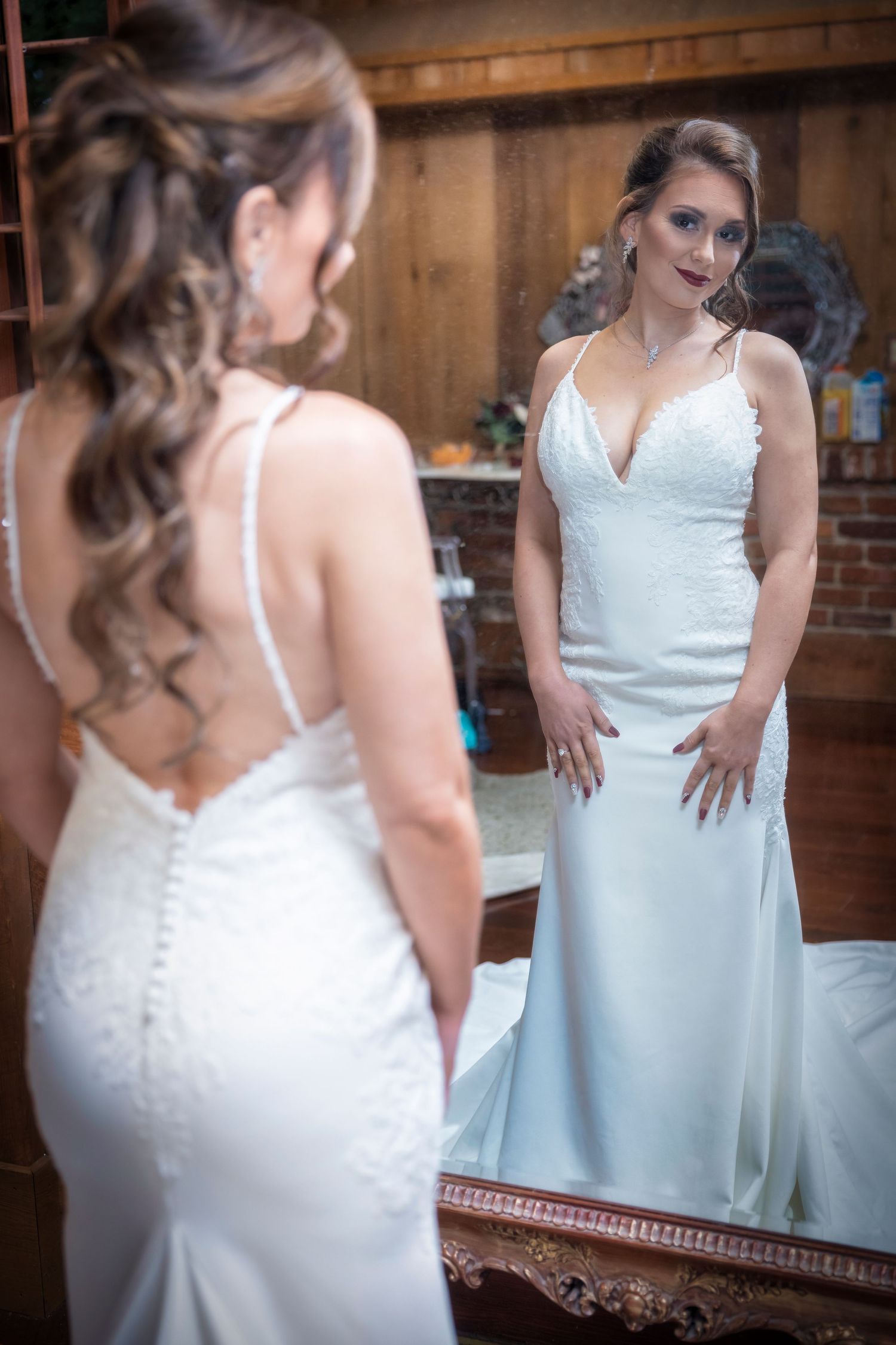 A bride in an elegant white fitted wedding gown with spaghetti straps looks at her reflection in an ornate mirror.