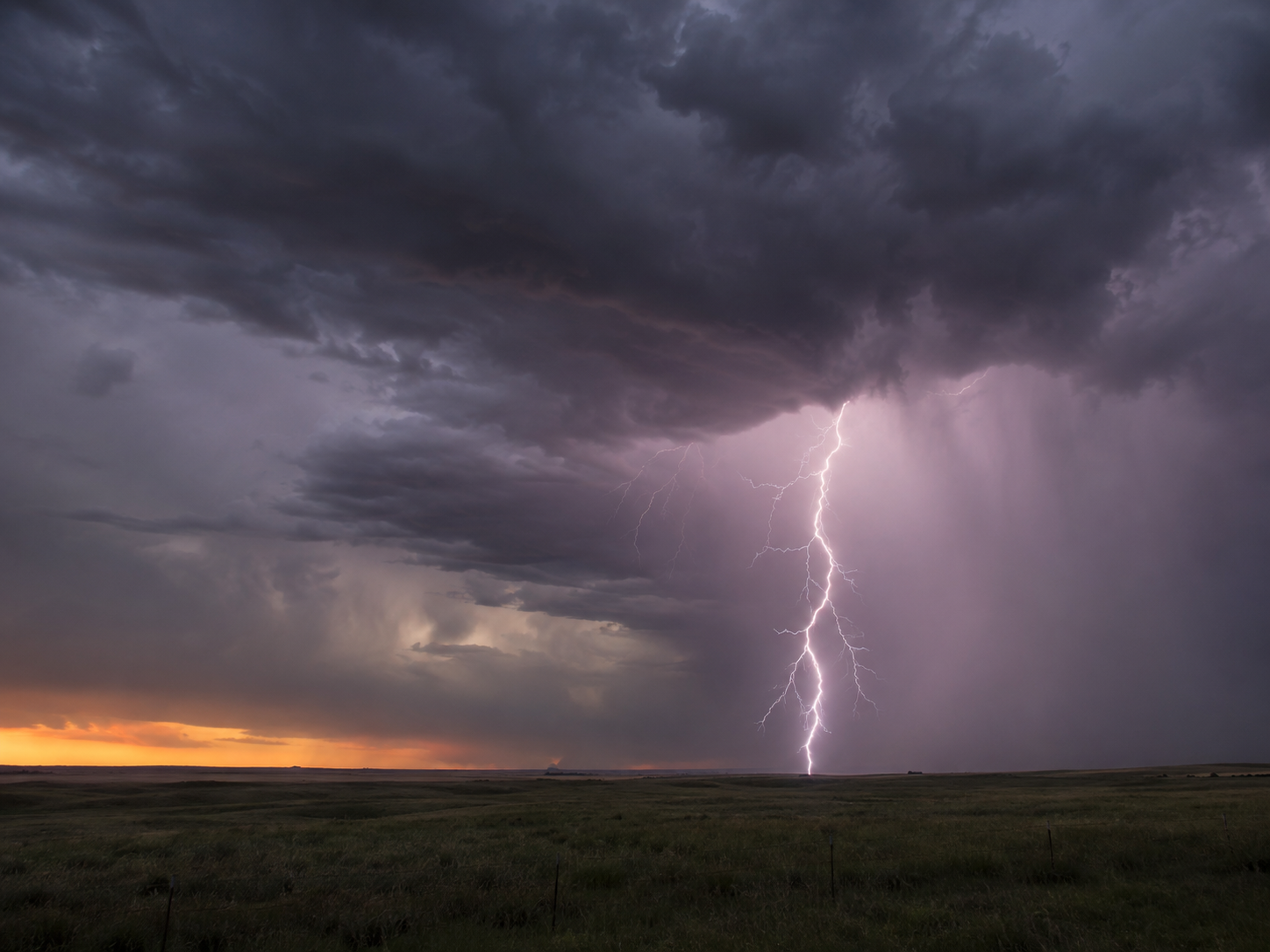 Dramatic lightning bolt illuminates storm clouds over a vast open prairie at sunset.