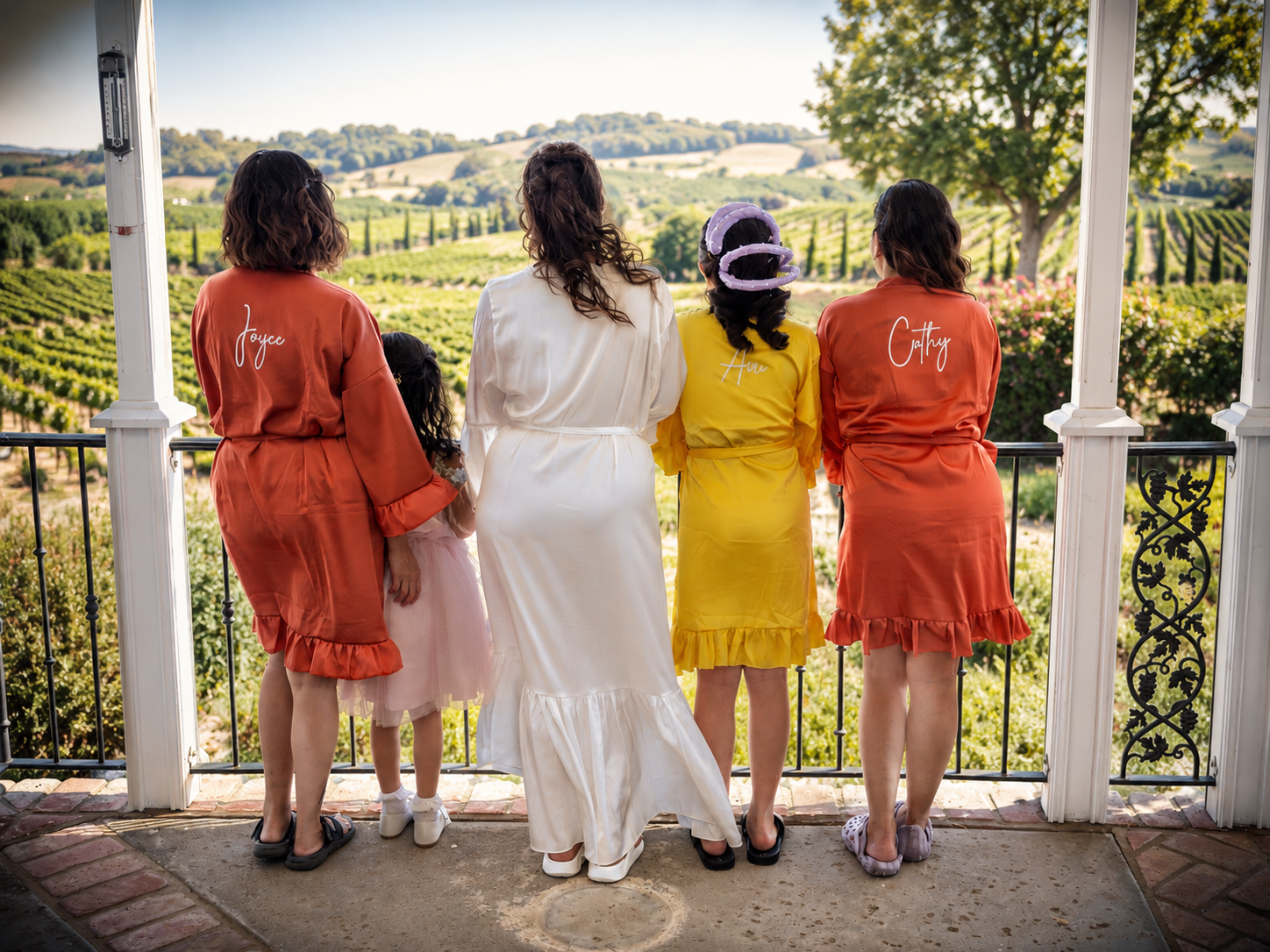 Bride and bridesmaids in colorful robes stand on a balcony overlooking a scenic vineyard landscape.
