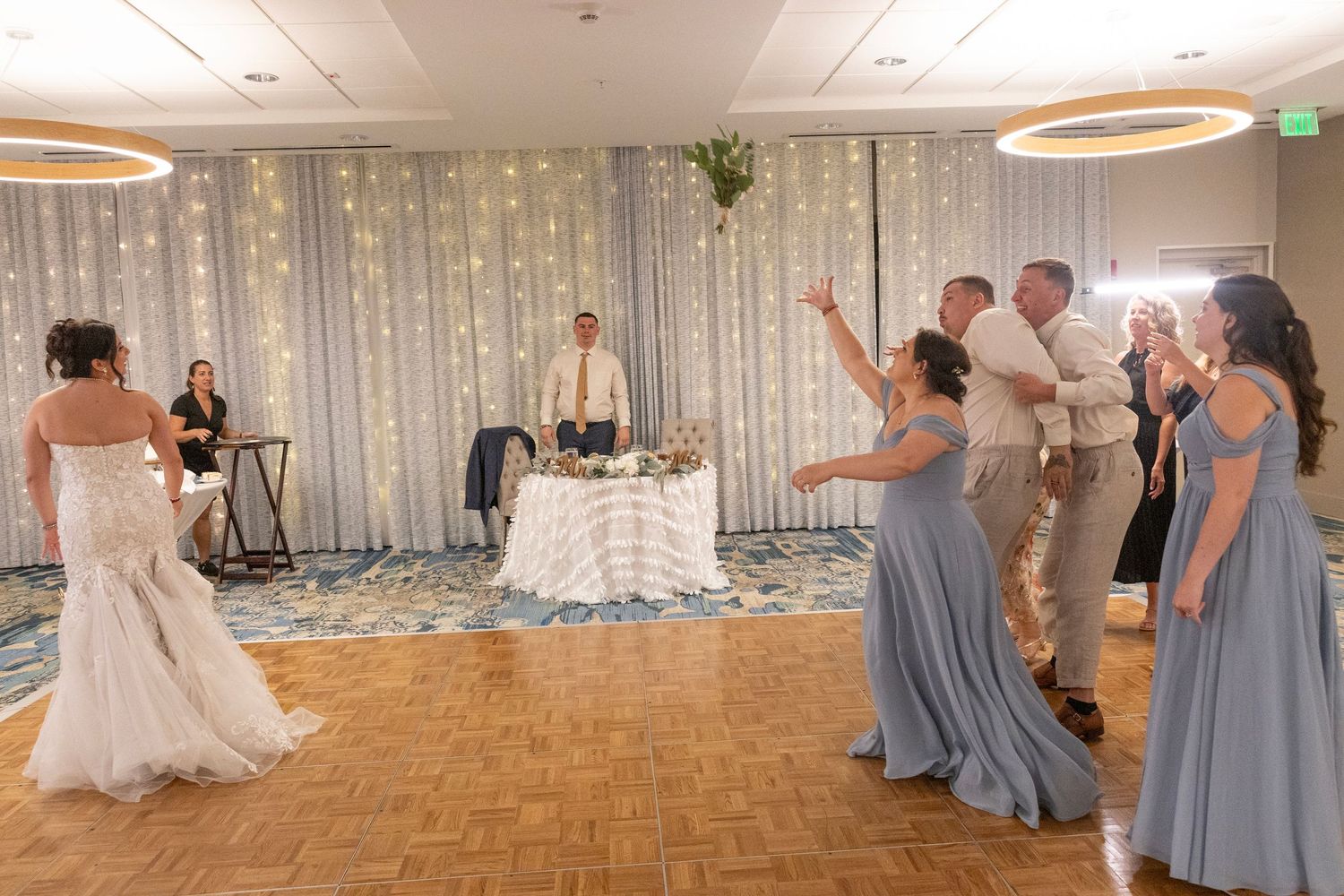 Bride tossing bouquet to bridesmaids in blue dresses at elegant wedding reception with fairy light backdrop.