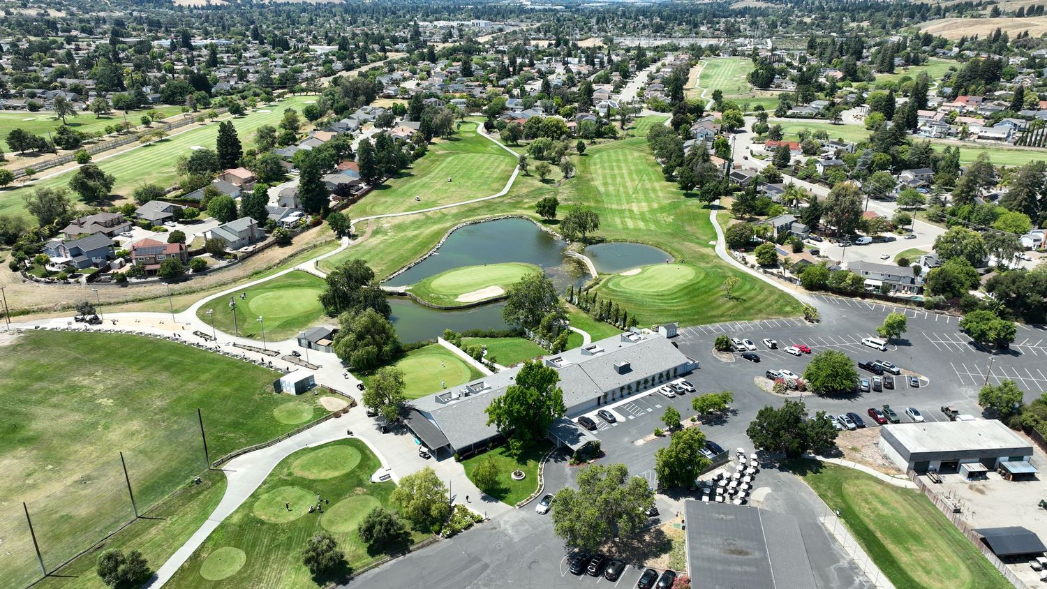 Aerial view of a lush green golf course with a pond, clubhouse, and parking lot surrounded by residential neighborhood.