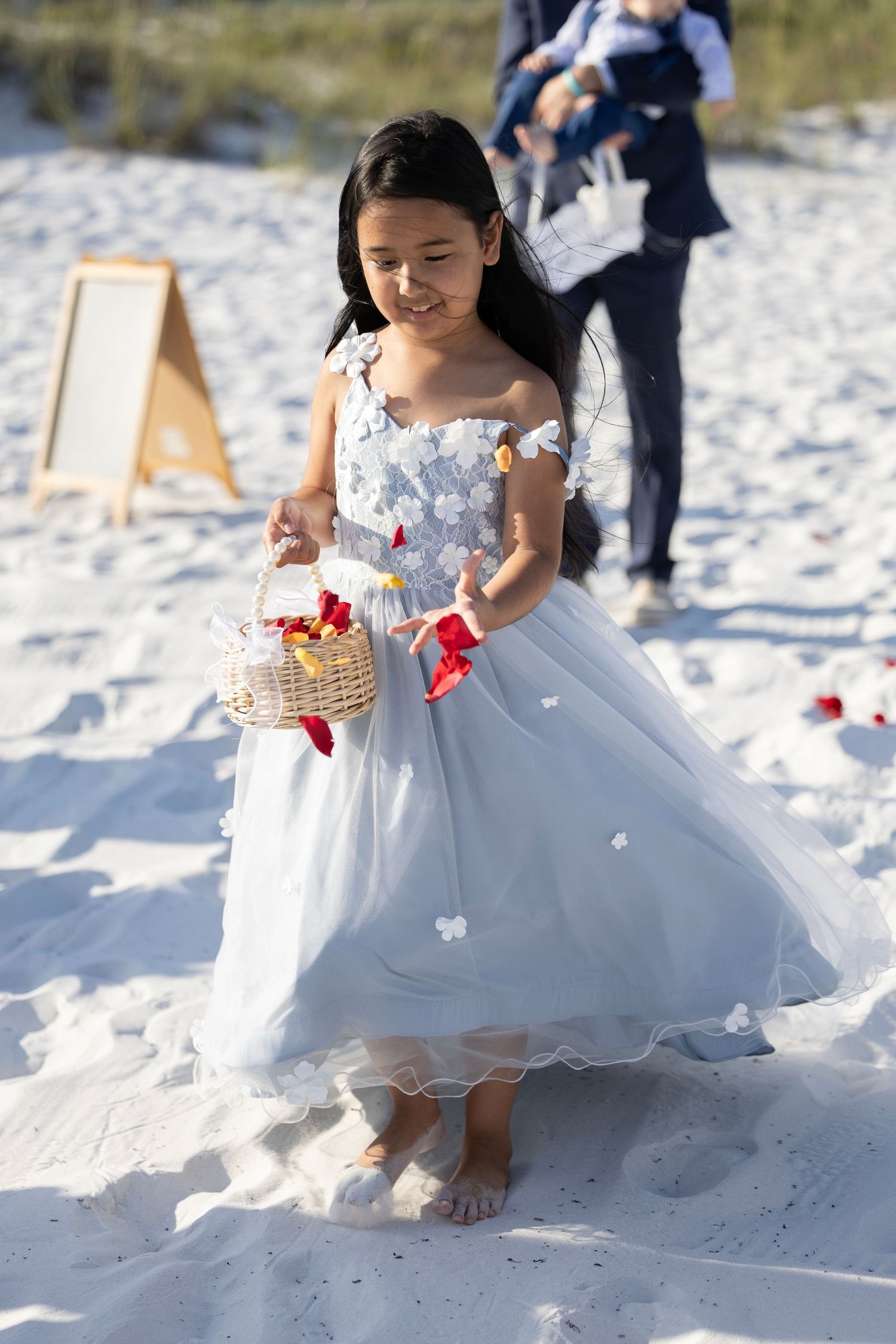 Young flower girl in white dress scattering rose petals on sandy beach during outdoor wedding ceremony.