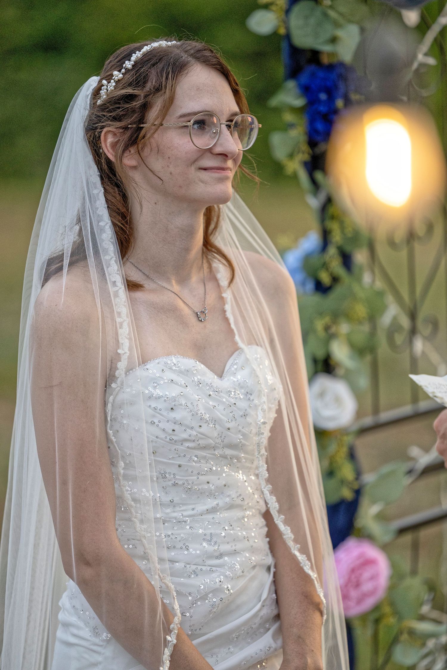 Smiling bride in a beaded strapless wedding gown and veil, wearing glasses, standing outdoors near a floral arch.
