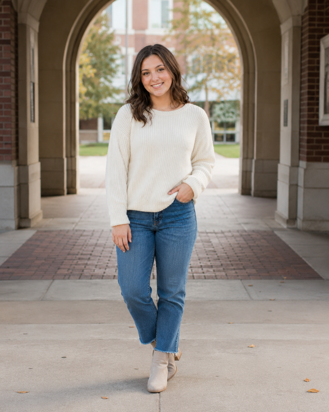 Smiling young woman in cream sweater and jeans walking through university archway.
