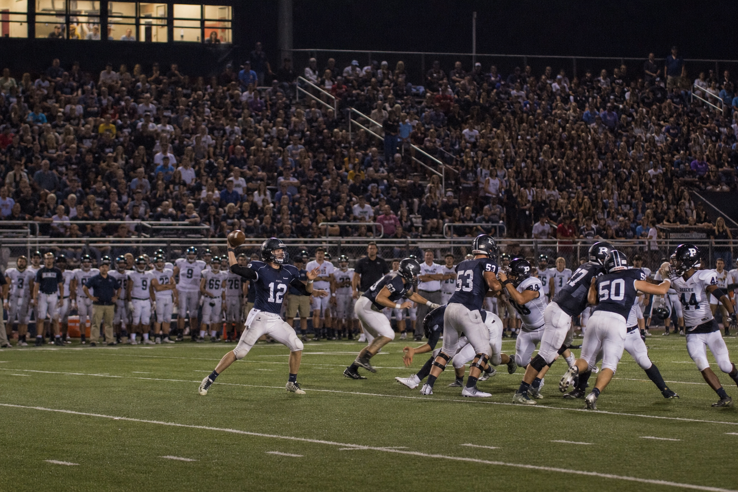 Quarterback #12 prepares to throw a pass during a packed night football game at a high school stadium.