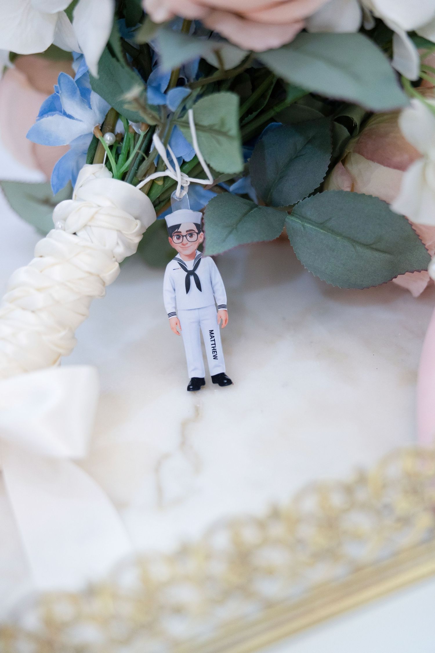 Miniature sailor figurine charm hanging from a wedding bouquet with pink and blue flowers on white fabric.