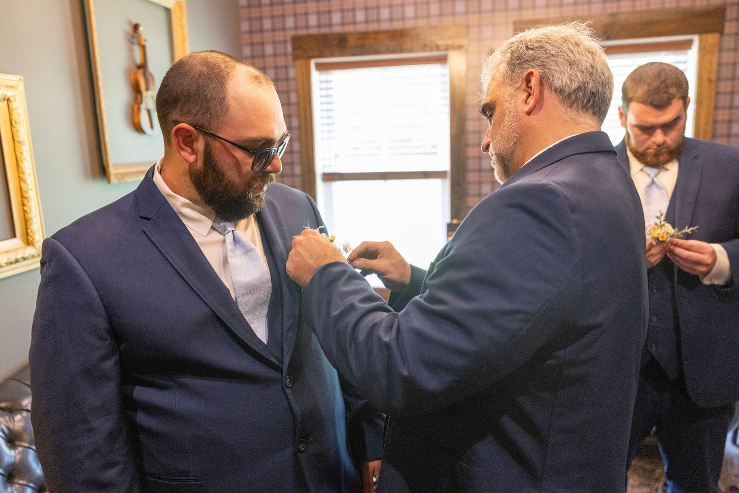 Men in navy suits helping each other with boutonnieres while getting ready for a wedding ceremony.