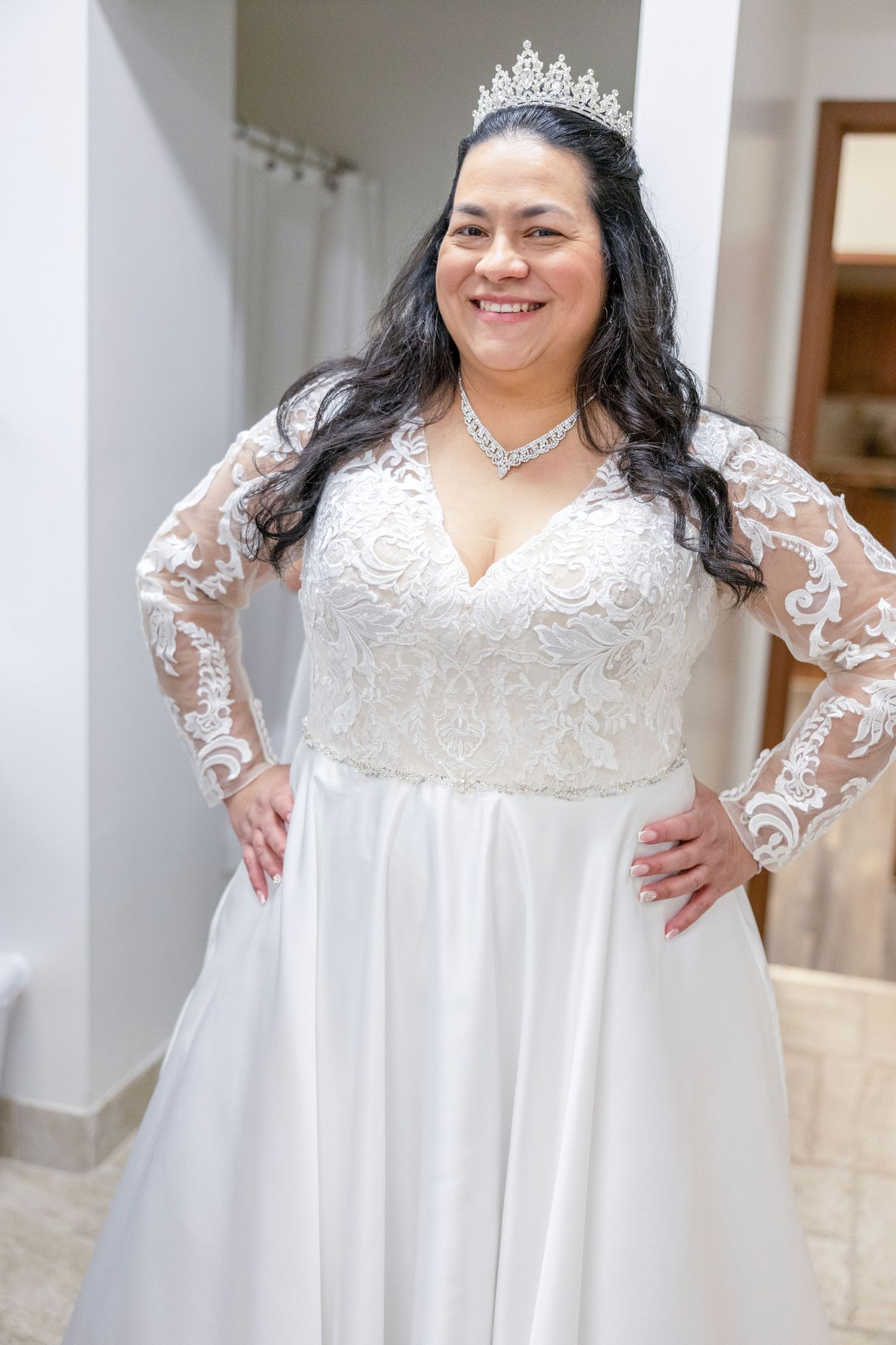 Plus-size bride wearing a white lace long-sleeve wedding gown with a crystal tiara and necklace, smiling with hands on hips.
