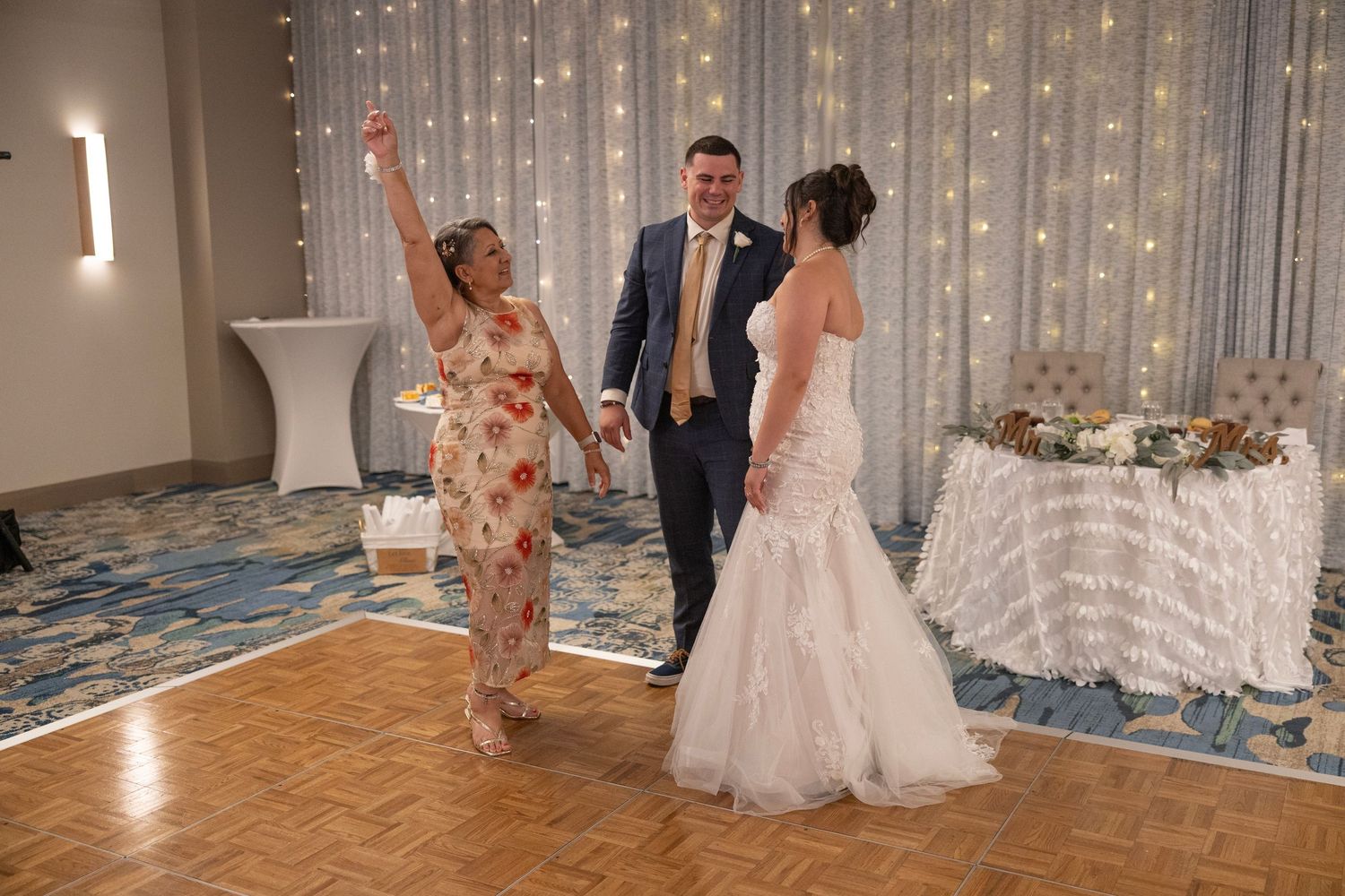 Bride in white mermaid gown dances with groom and guest at elegant wedding reception with fairy lights.